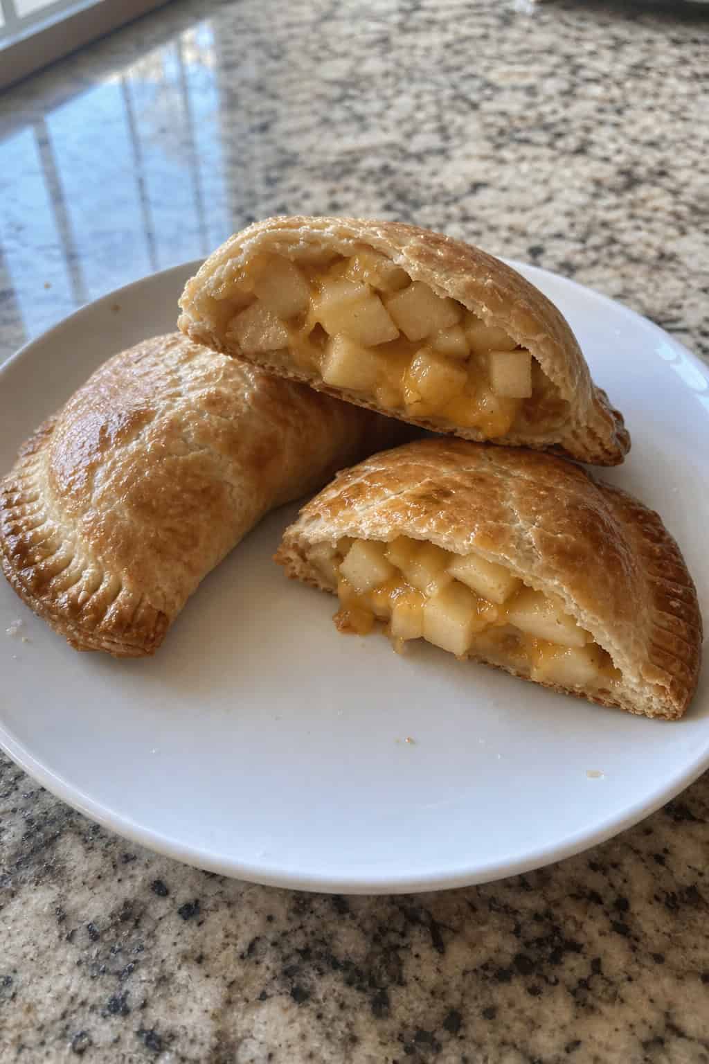 A wooden board with golden-brown apple-cheddar maple hand pies, a bowl of maple syrup, apple slices, and cheddar cheese pieces on a kitchen counter.