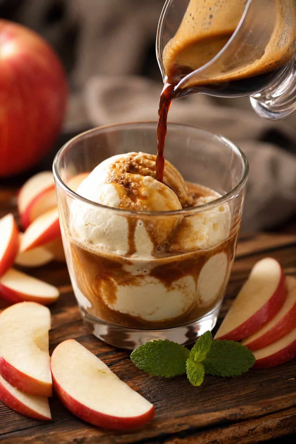 A glass cup of vanilla ice cream with espresso being poured over it, surrounded by sliced red apples and a mint garnish on a wooden surface.