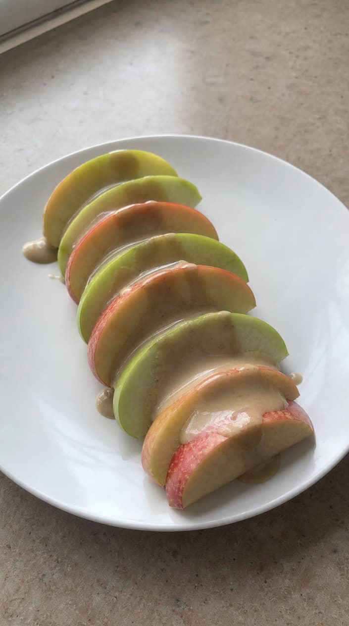 Fresh apple slices drizzled with tahini sauce on a wooden cutting board with a small bowl of tahini and whole apples in the background.