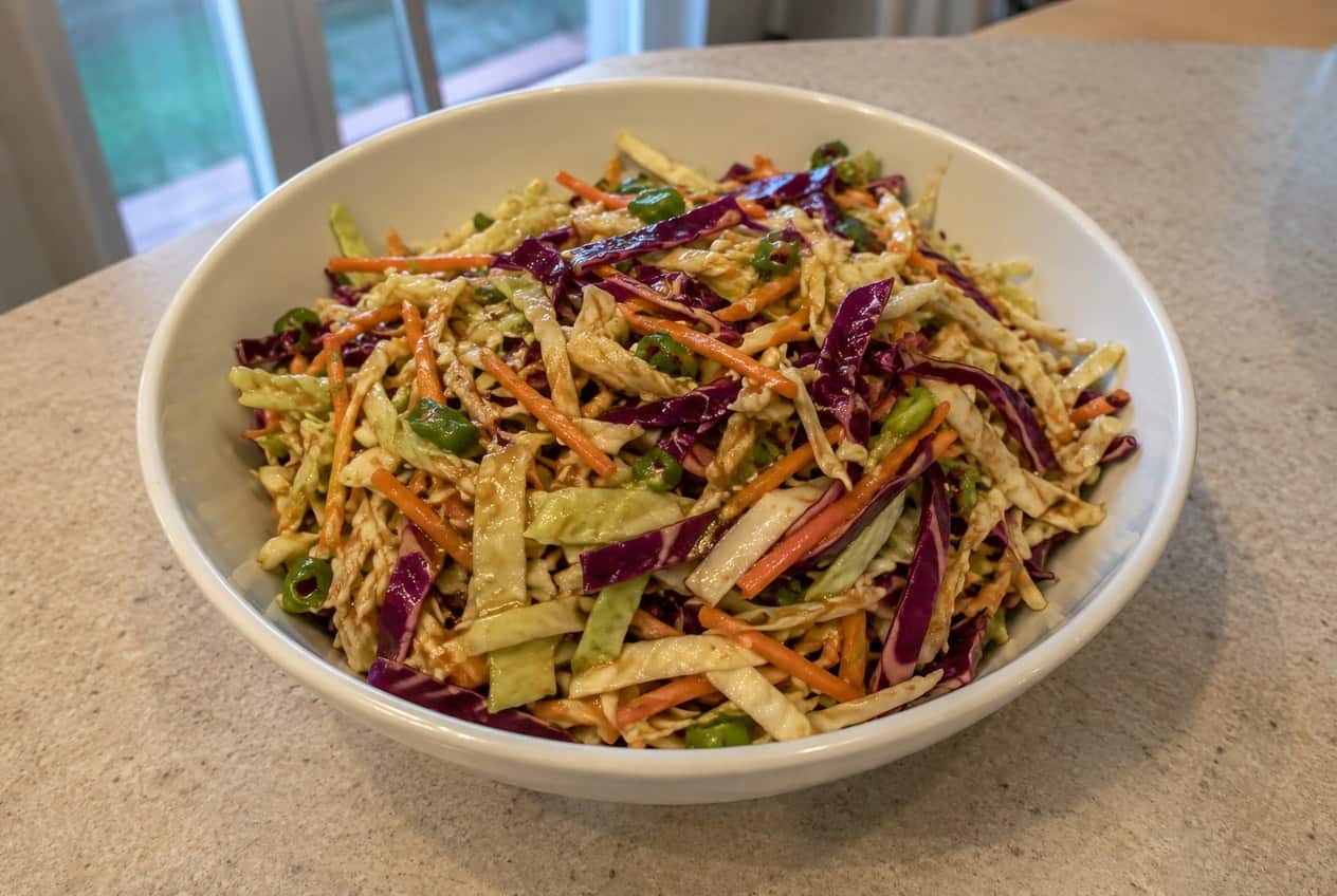 A deep bowl of shredded green and purple cabbage slaw flecked with orange carrot ribbons, sliced green onions, and cilantro, scattered with white sesame seeds, on a wooden surface.