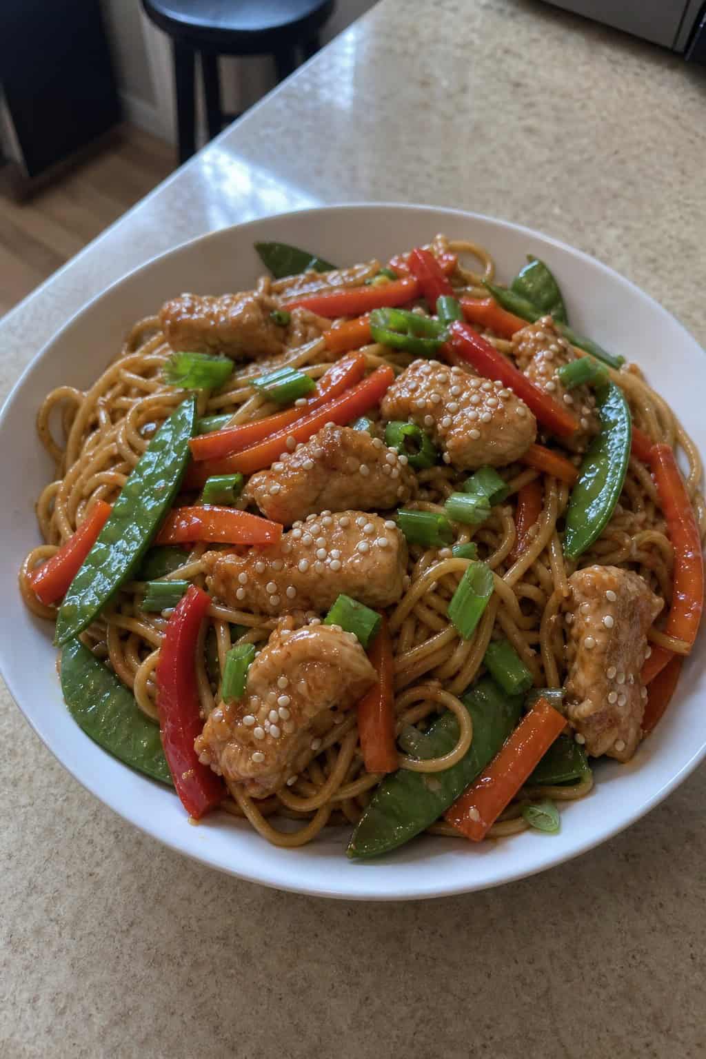 A deep bowl of lo mein noodles tossed with shredded chicken, shredded carrots, and snow peas in a glossy sesame-soy sauce, topped with sliced green onions and sesame seeds.