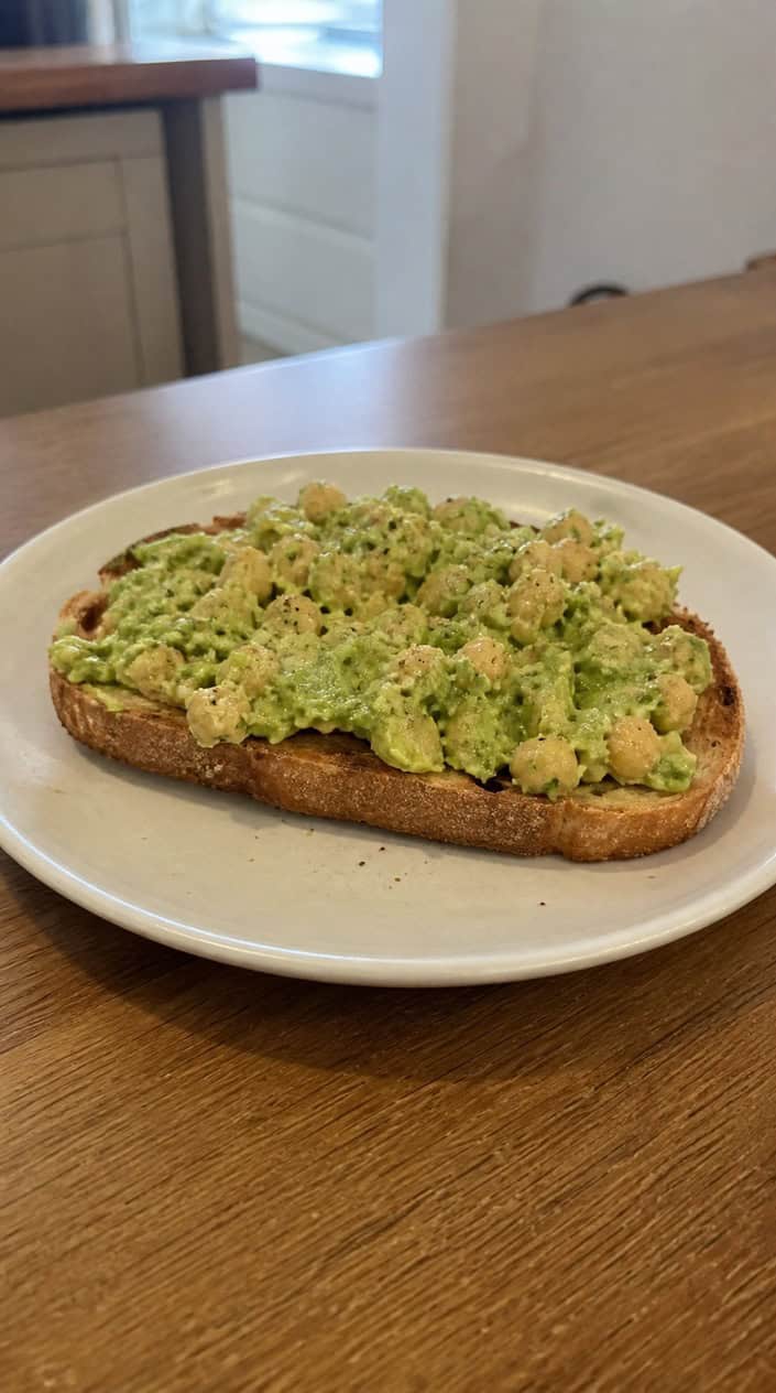 A slice of toasted sourdough topped with avocado chickpea smash garnished with herbs on a wooden board.