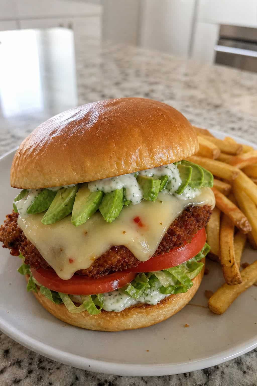 A grilled chicken sandwich with sliced avocado, melted pepper jack cheese, lettuce, and tomato on a toasted bun, served with fries and ranch dressing on a wooden board