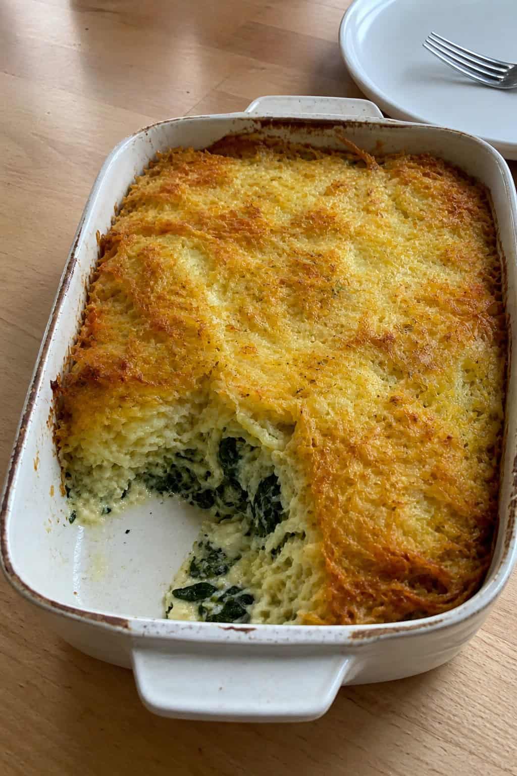 A baked casserole dish filled with baby spinach, ricotta cheese, and crispy hashbrowns on a wooden table with a serving spatula nearby.