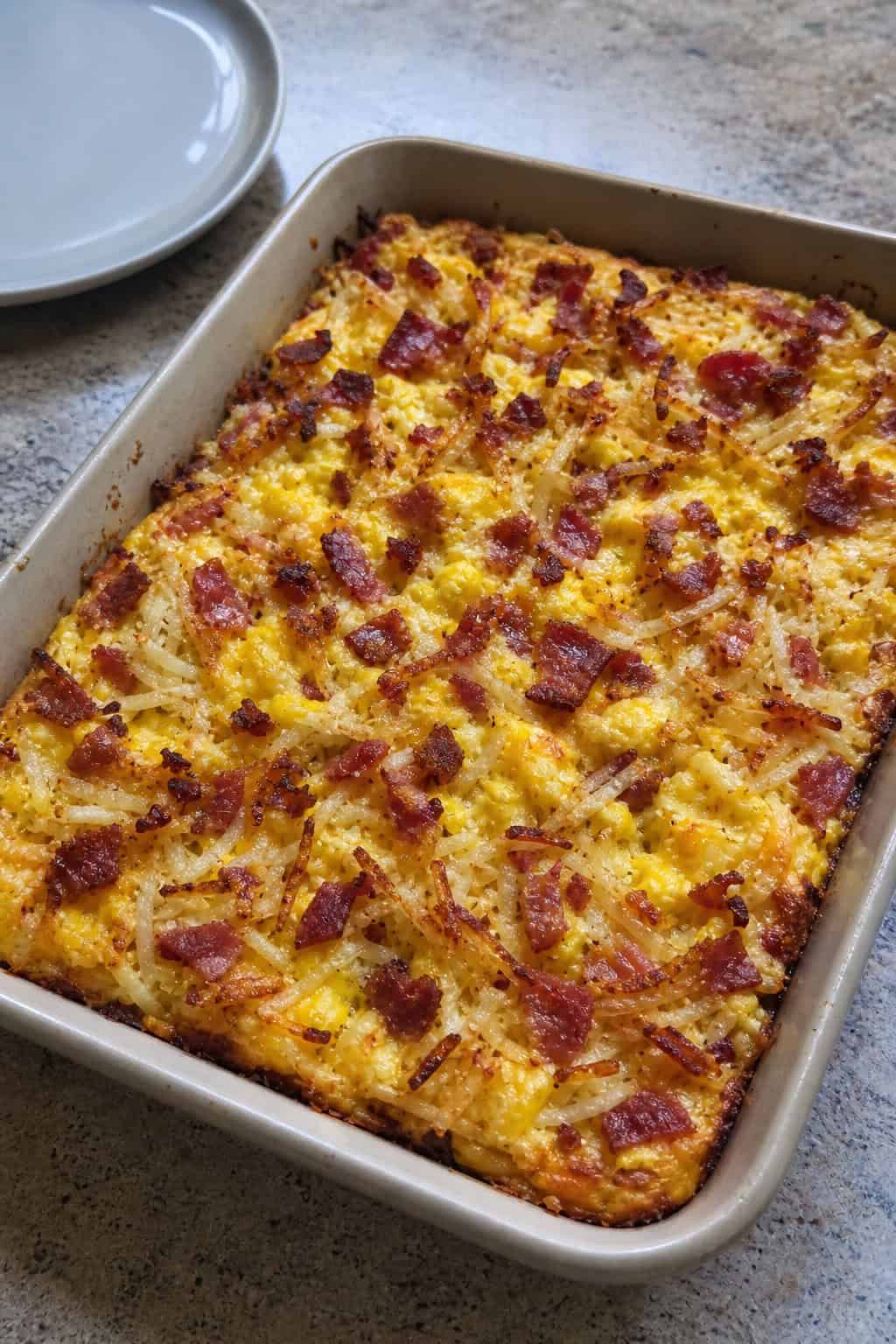 A baked breakfast casserole with crispy bacon pieces, egg custard, and shredded hashbrowns in a ceramic dish on a wood table with a fork and orange juice glass nearby.