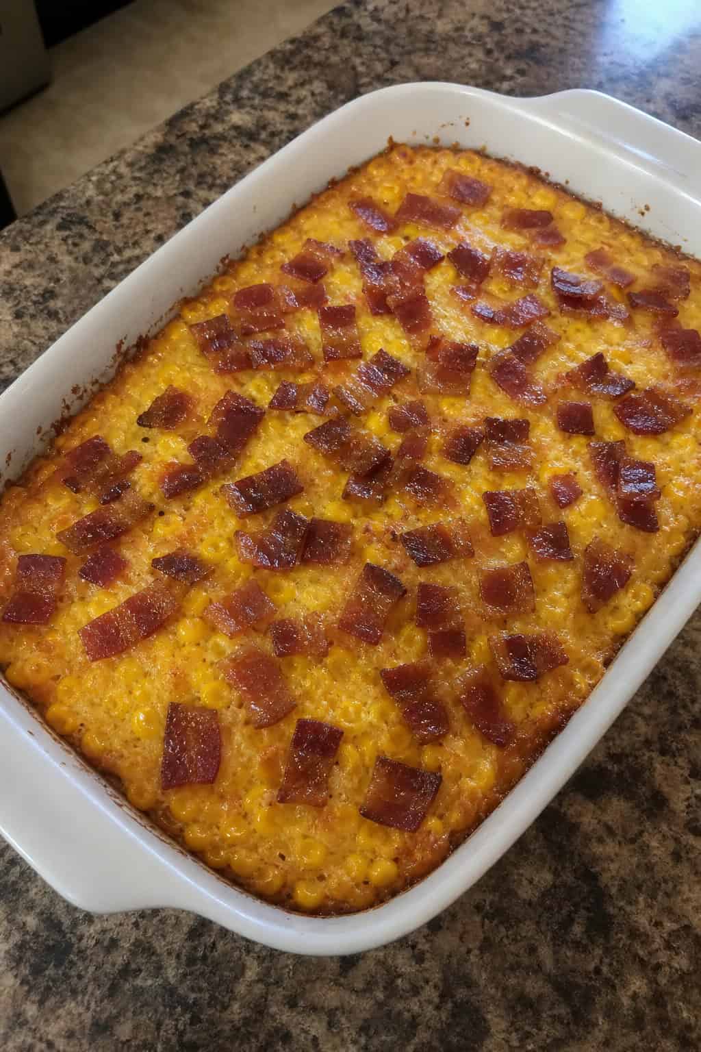 A bubbling corn casserole topped with a full layer of crispy bacon strips in a rectangular baking dish on a wooden table, with a bowl of shredded cheese and a pepper in the background.