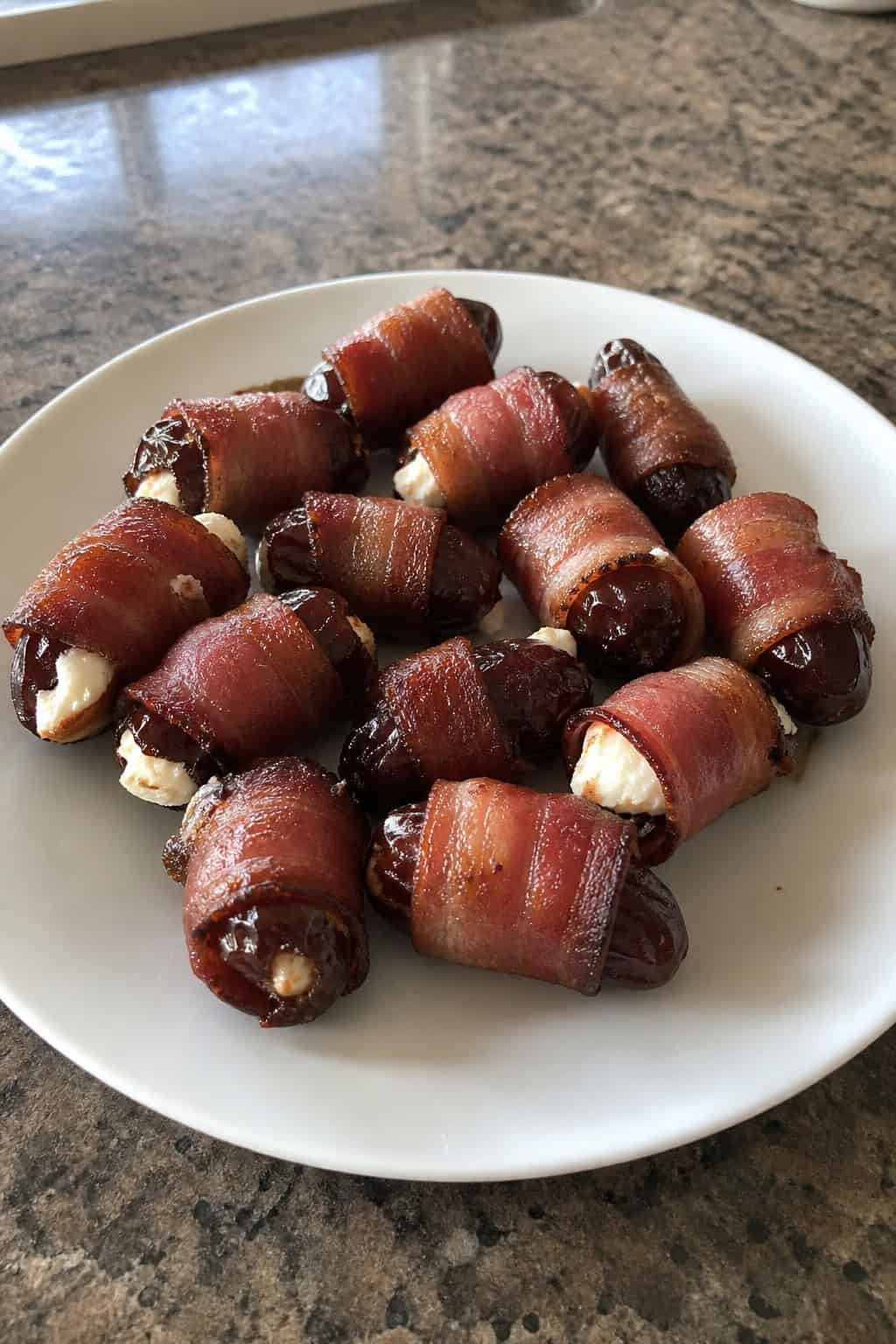 A platter of bacon-wrapped dates stuffed with goat cheese, garnished with fresh herbs on a wooden board.