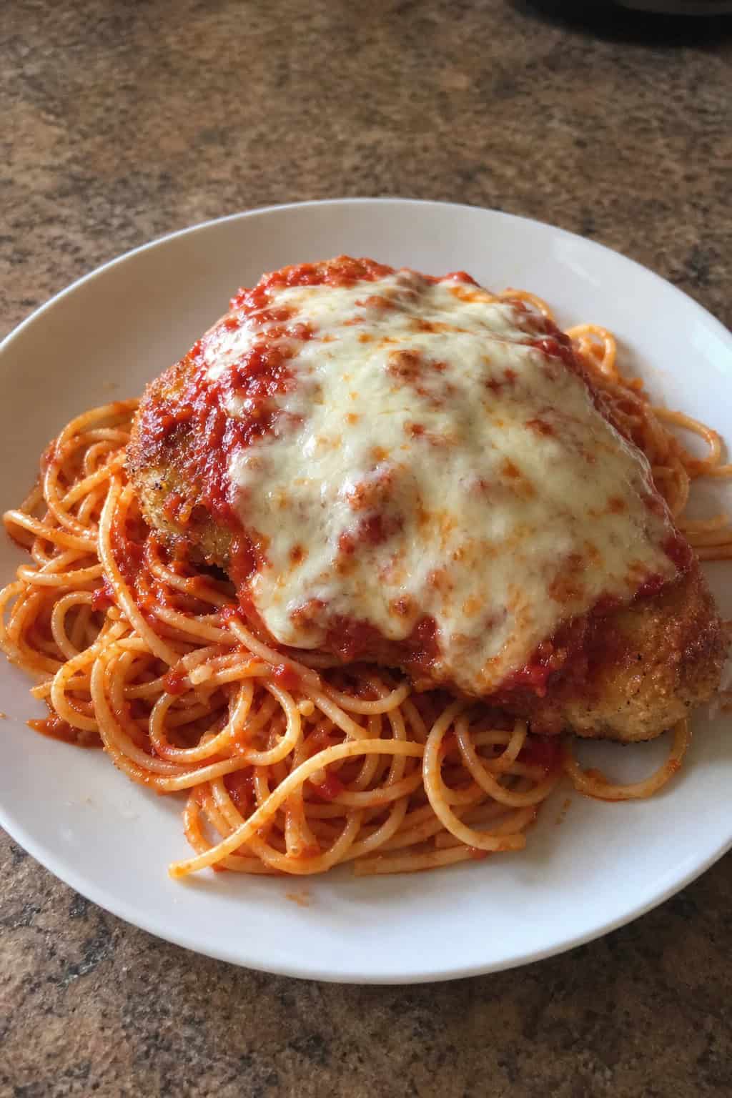 A plate of baked chicken parmesan with melted cheese and marinara sauce served over spaghetti pasta, garnished with basil and Parmesan cheese.