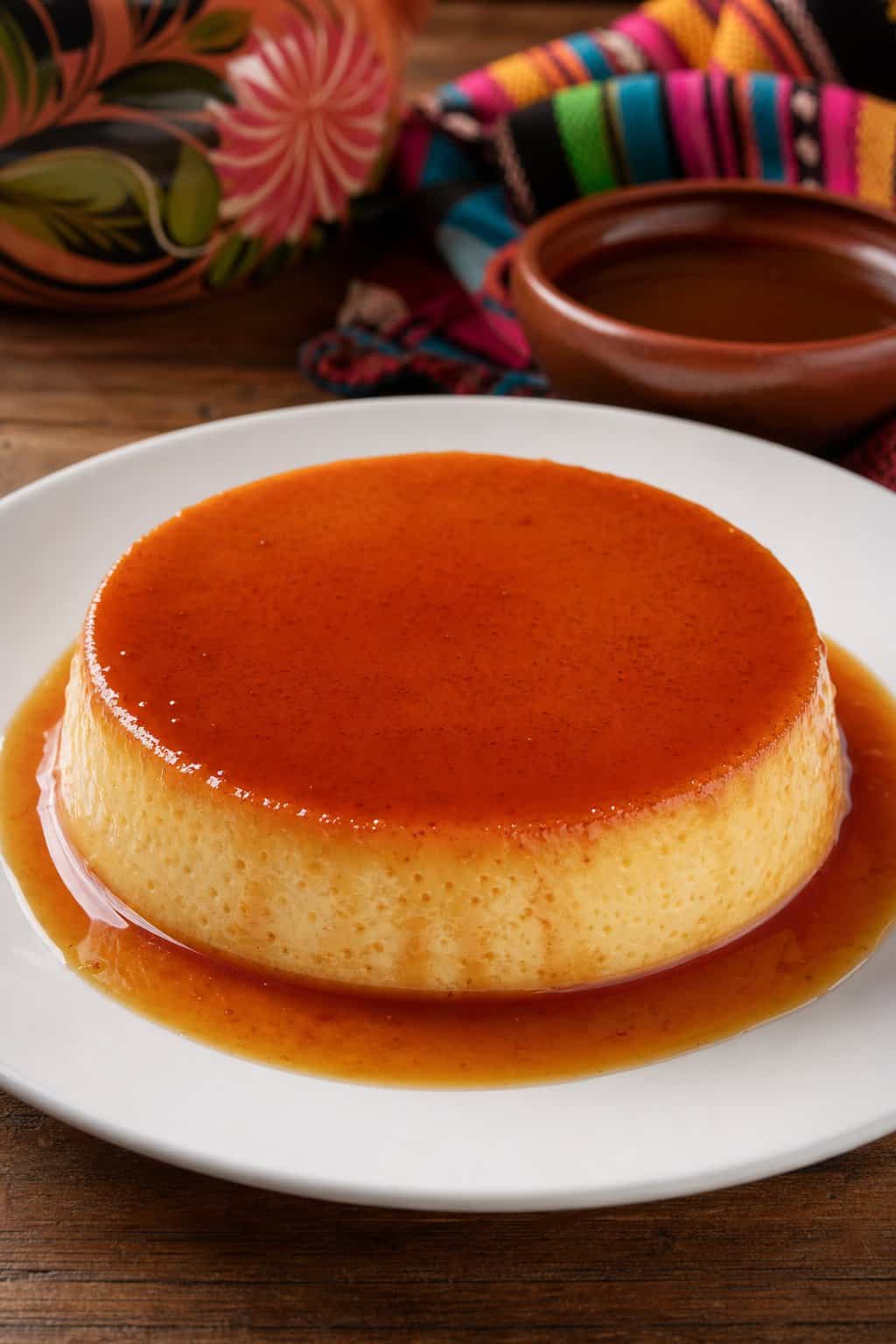 A plate with a baked flan topped with caramel sauce on a wooden table with a colorful cloth in the background.
