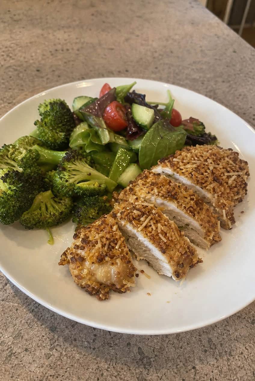 A plate of baked Parmesan crusted chicken breasts with a golden, textured coating, garnished with fresh flat-leaf parsley on a rustic wooden table.