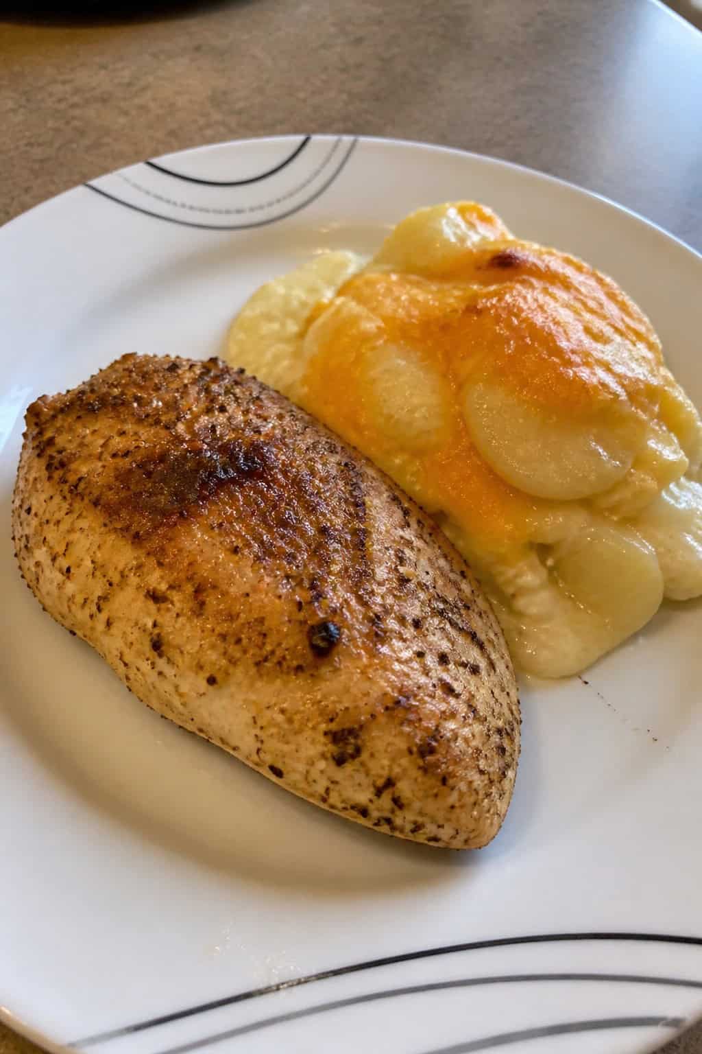 A plate with baked ranch chicken breast and cheesy potatoes garnished with herbs on a wooden table.