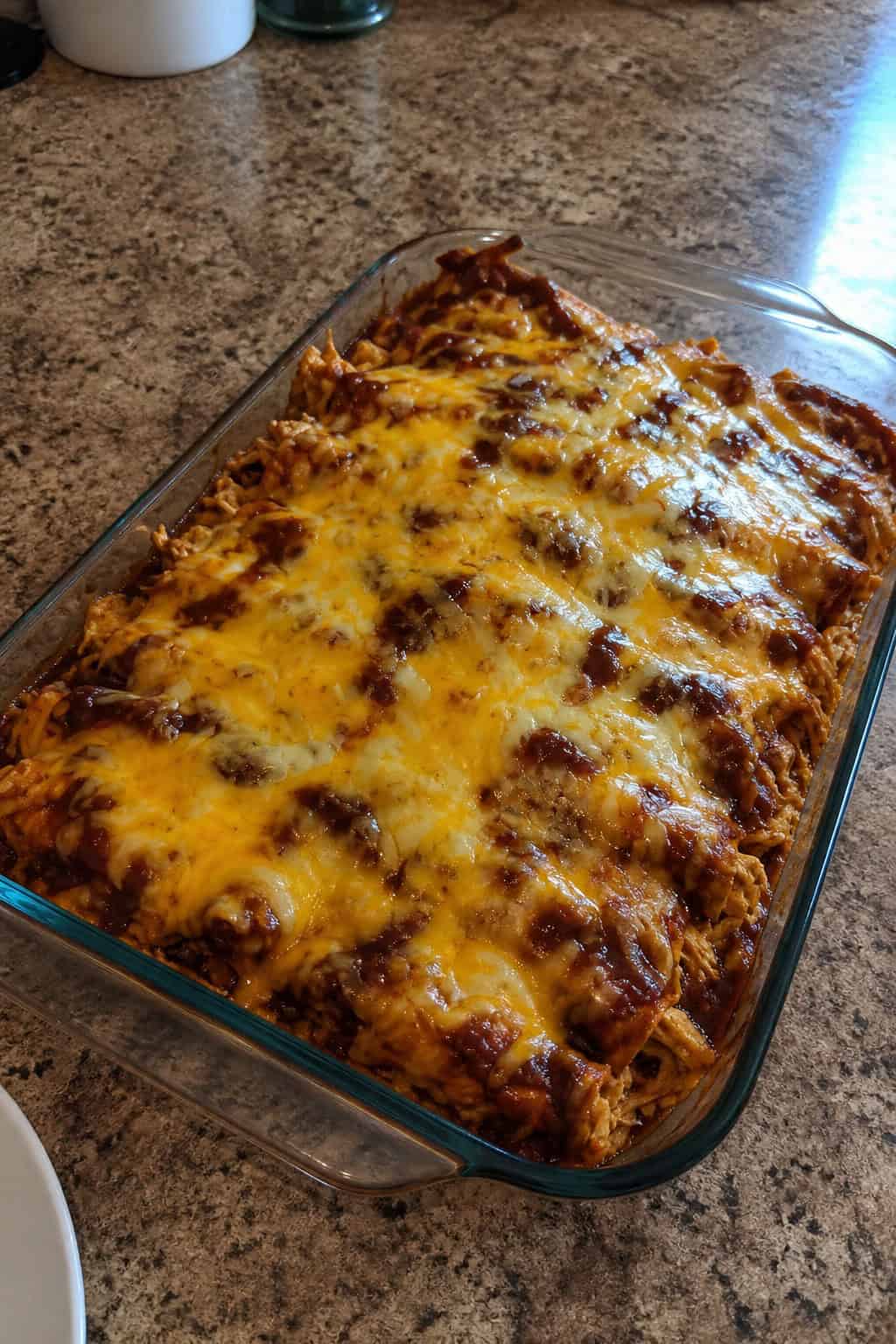 A freshly baked BBQ chicken enchilada casserole in a ceramic dish with caramelized edges, topped with sliced green onions and fresh cilantro, with a jar of BBQ sauce and a bowl of tortillas visible nearby on the wooden table.