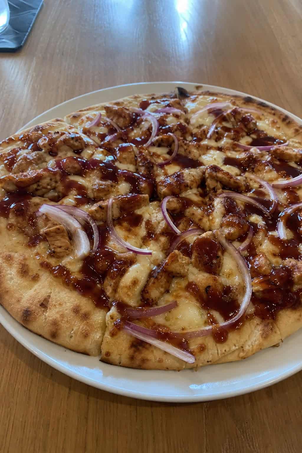 A naan-based BBQ chicken pizza on a wooden board, topped with melted mozzarella and cheddar, thinly sliced red onion rings, and scattered fresh cilantro leaves.