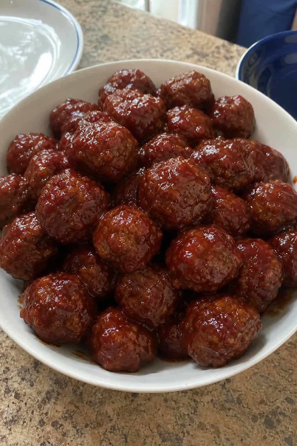 A bowl of BBQ meatballs coated in sauce with parsley garnish on a wooden table, accompanied by small bowls of sauce and grapes.