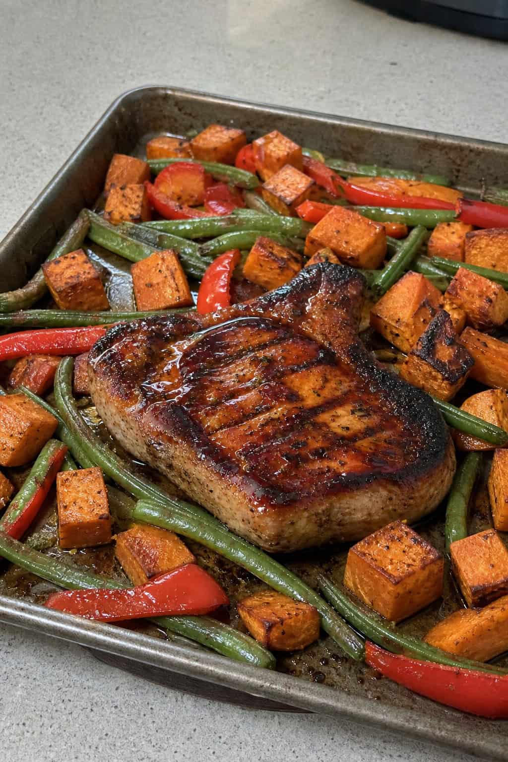 A thick bone-in pork chop glazed with dark BBQ sauce resting beside cubed roasted sweet potatoes and colorful mixed vegetables on a foil-lined rimmed baking sheet.