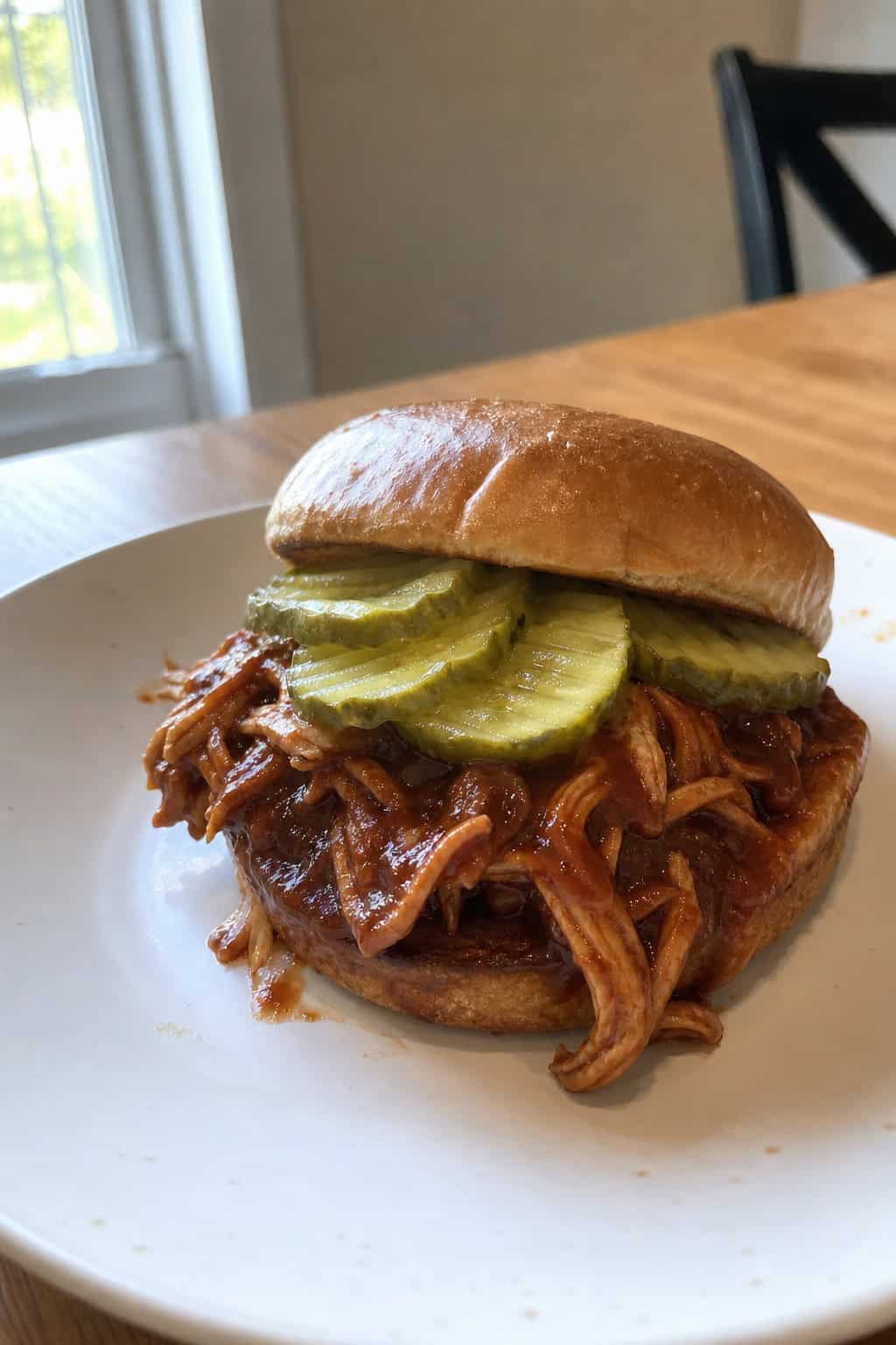 A BBQ pulled chicken sandwich stacked high on a wooden board with a small bowl of BBQ sauce and a pile of pickle chips nearby