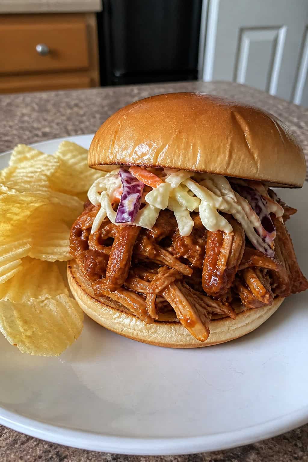 A pulled chicken sandwich with barbecue sauce and coleslaw on a toasted bun, served with potato chips on a wooden table.