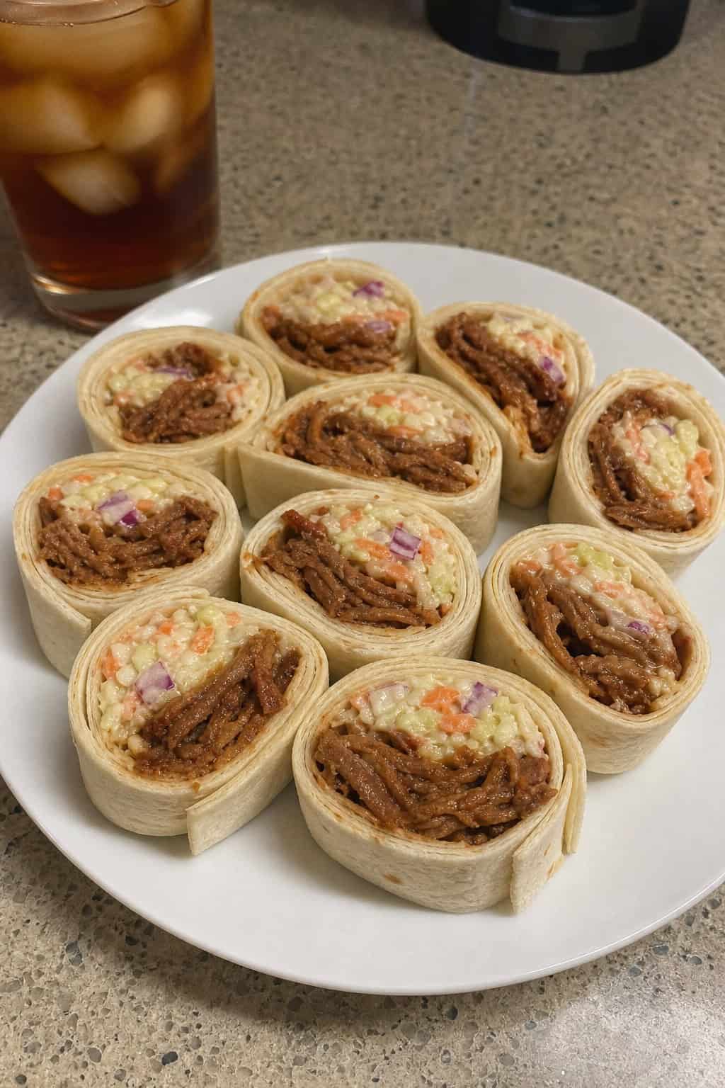 Plate of BBQ pulled pork and coleslaw pinwheel appetizers showing dark smoky pulled pork and pale shredded coleslaw inside sliced flour tortilla rolls on a wooden serving board.