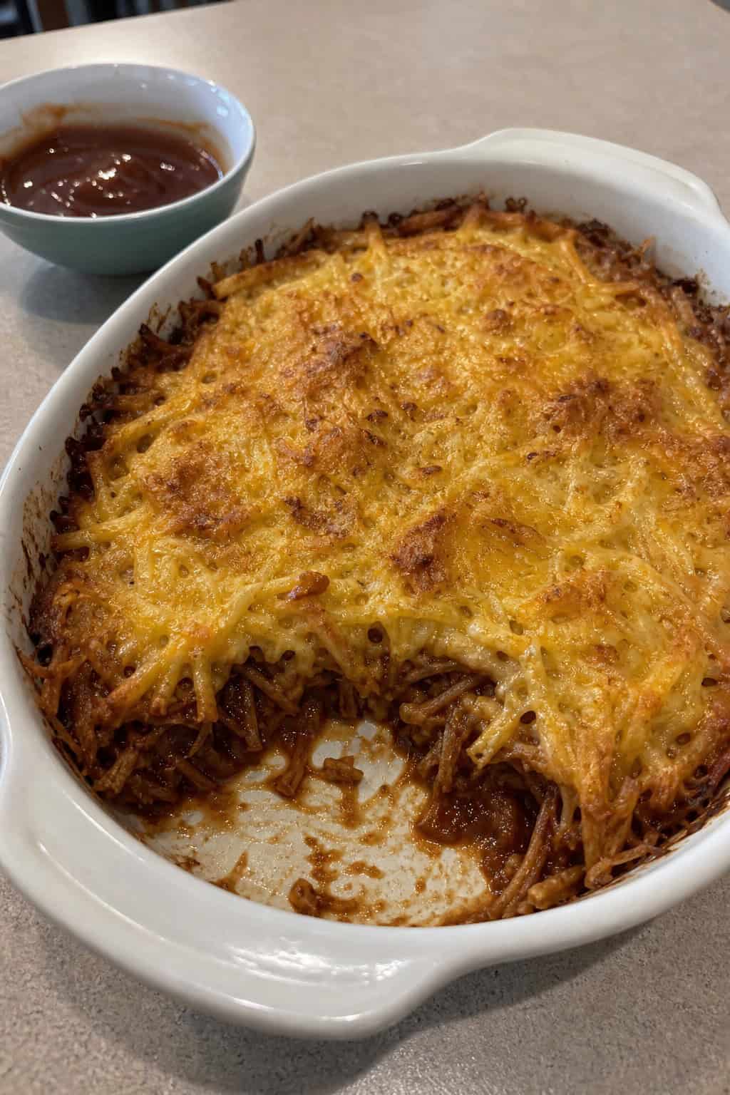 A baked BBQ pulled pork hashbrown casserole in a ceramic dish on a wooden table with a fork and barbecue sauce nearby.