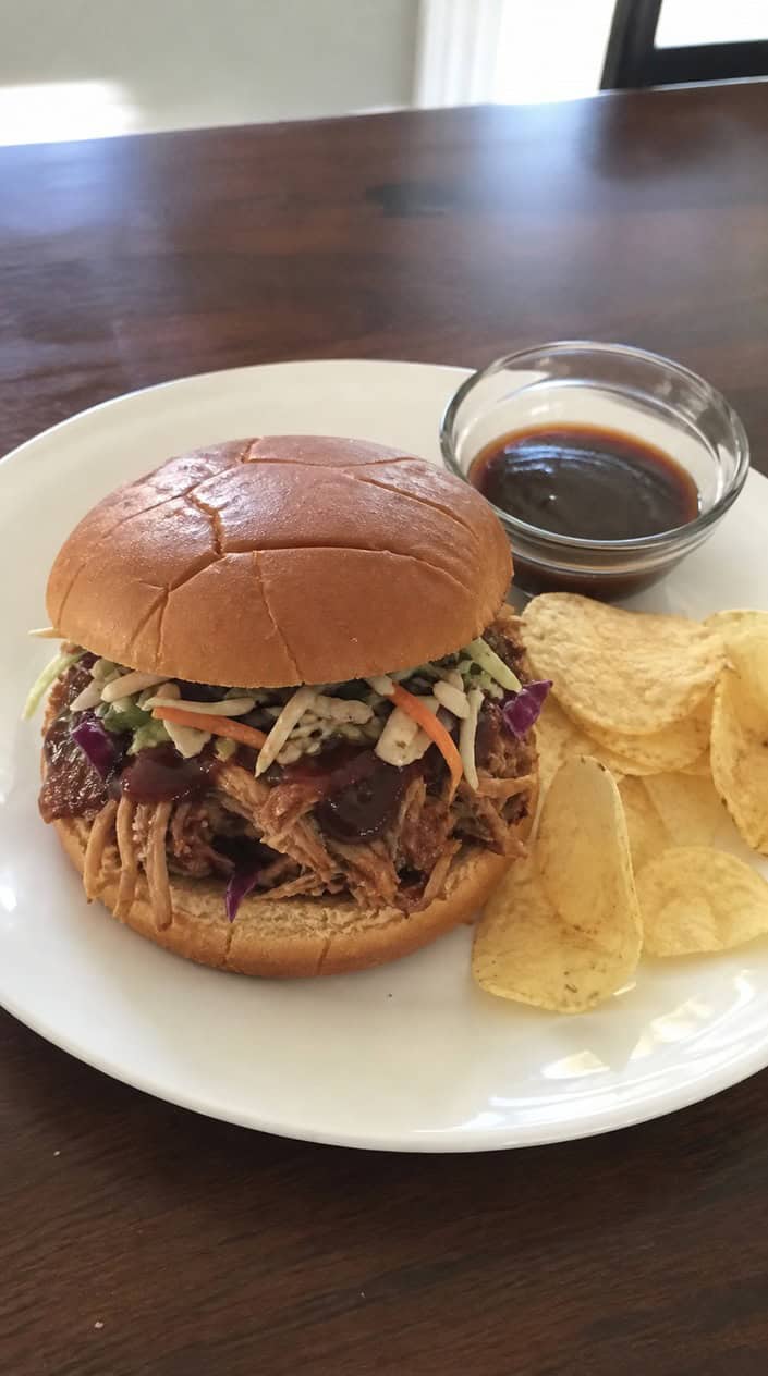 A pulled pork sloppy joe loaded with creamy coleslaw on a toasted brioche bun, served on a wooden board next to a small bowl of deep red barbecue sauce and a handful of ridged potato chips.