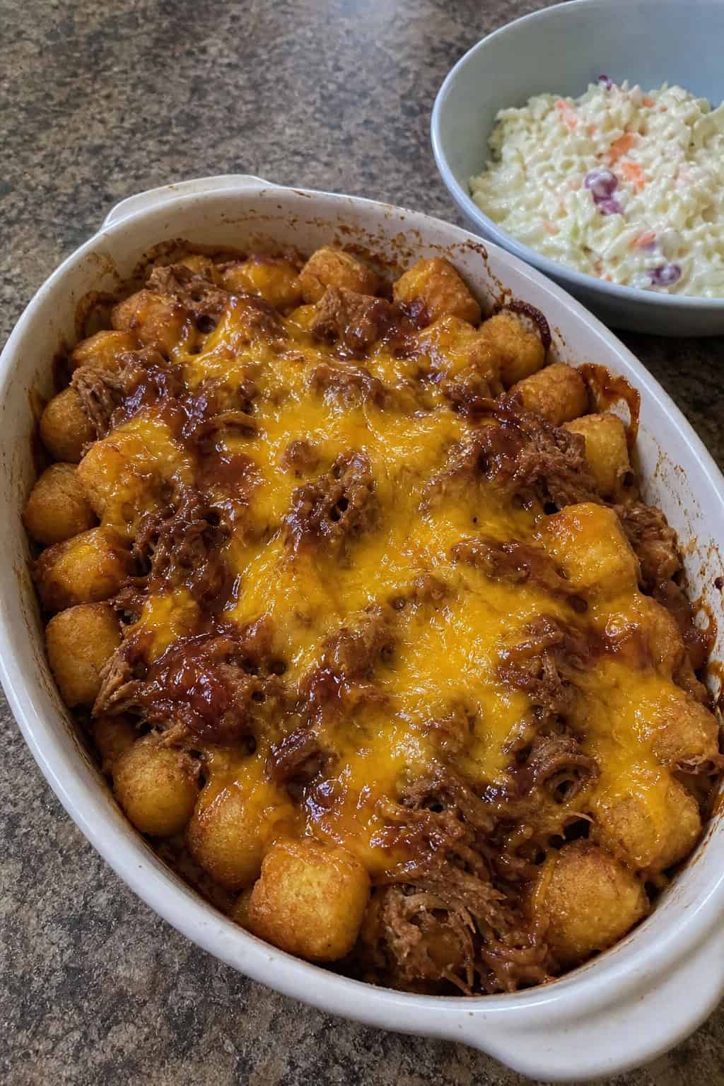 A BBQ pulled pork tater tot casserole in a rectangular baking dish with golden tots covering the top, shredded pork and melted cheddar visible at the edges, and a bowl of coleslaw beside it on a wooden table.