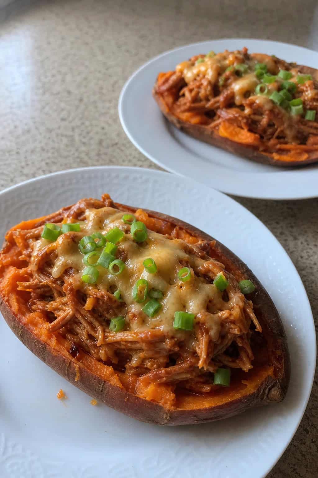 Two halved roasted sweet potatoes on a wooden table, their orange flesh topped with saucy BBQ shredded chicken, a scattering of shredded cheddar, and sliced green onions, with small bowls of BBQ sauce and fresh herbs nearby.