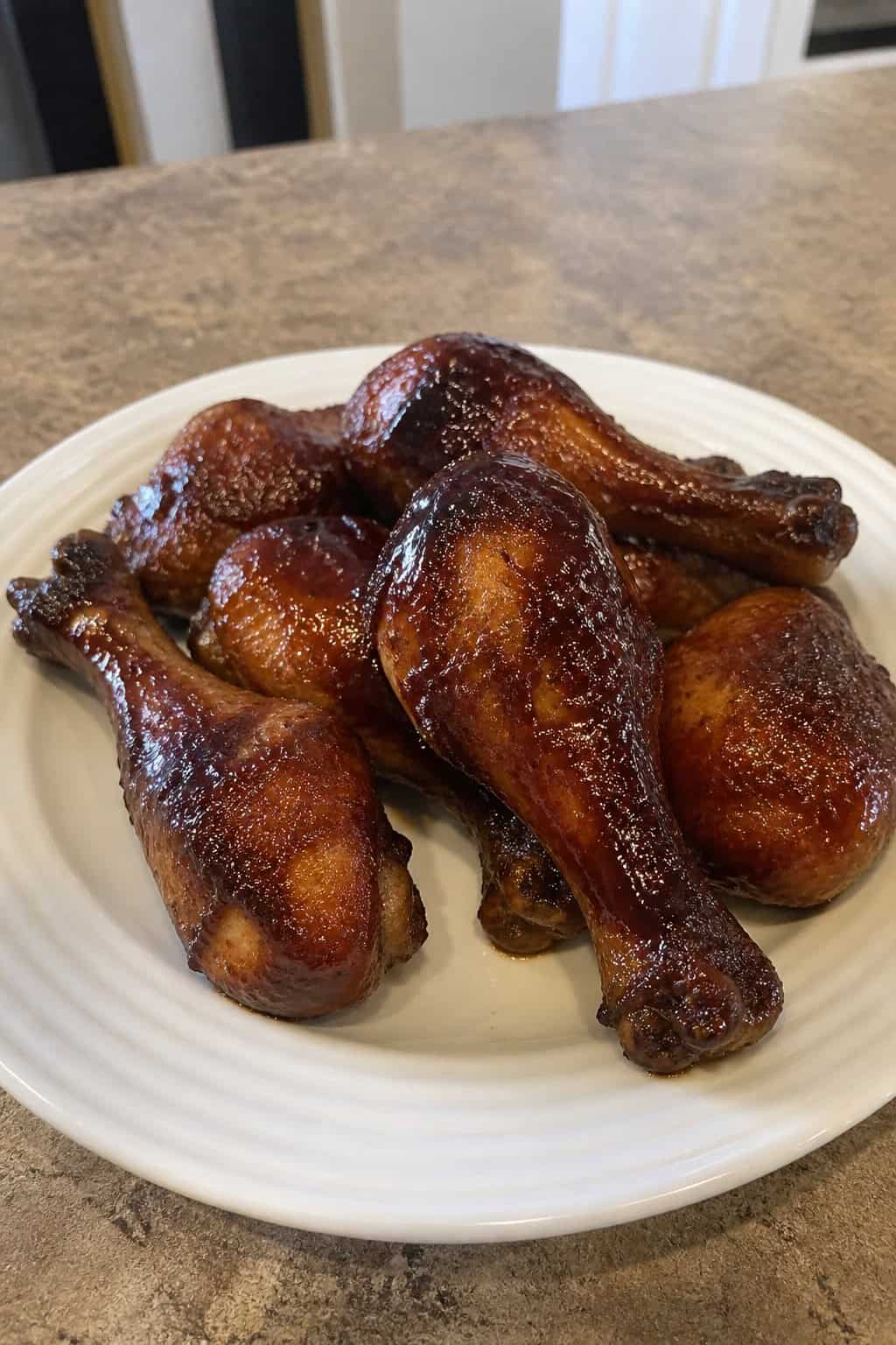 Deeply glazed BBQ chicken drumsticks piled on a round wooden plate with grilled vegetables and a scatter of fresh herbs alongside.
