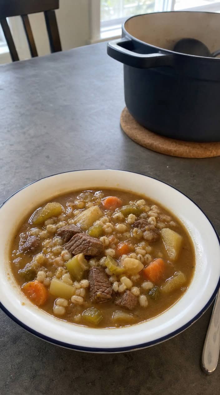 A bowl of beef and barley stew on a wooden table with a spoon and a freezer bag in the background.