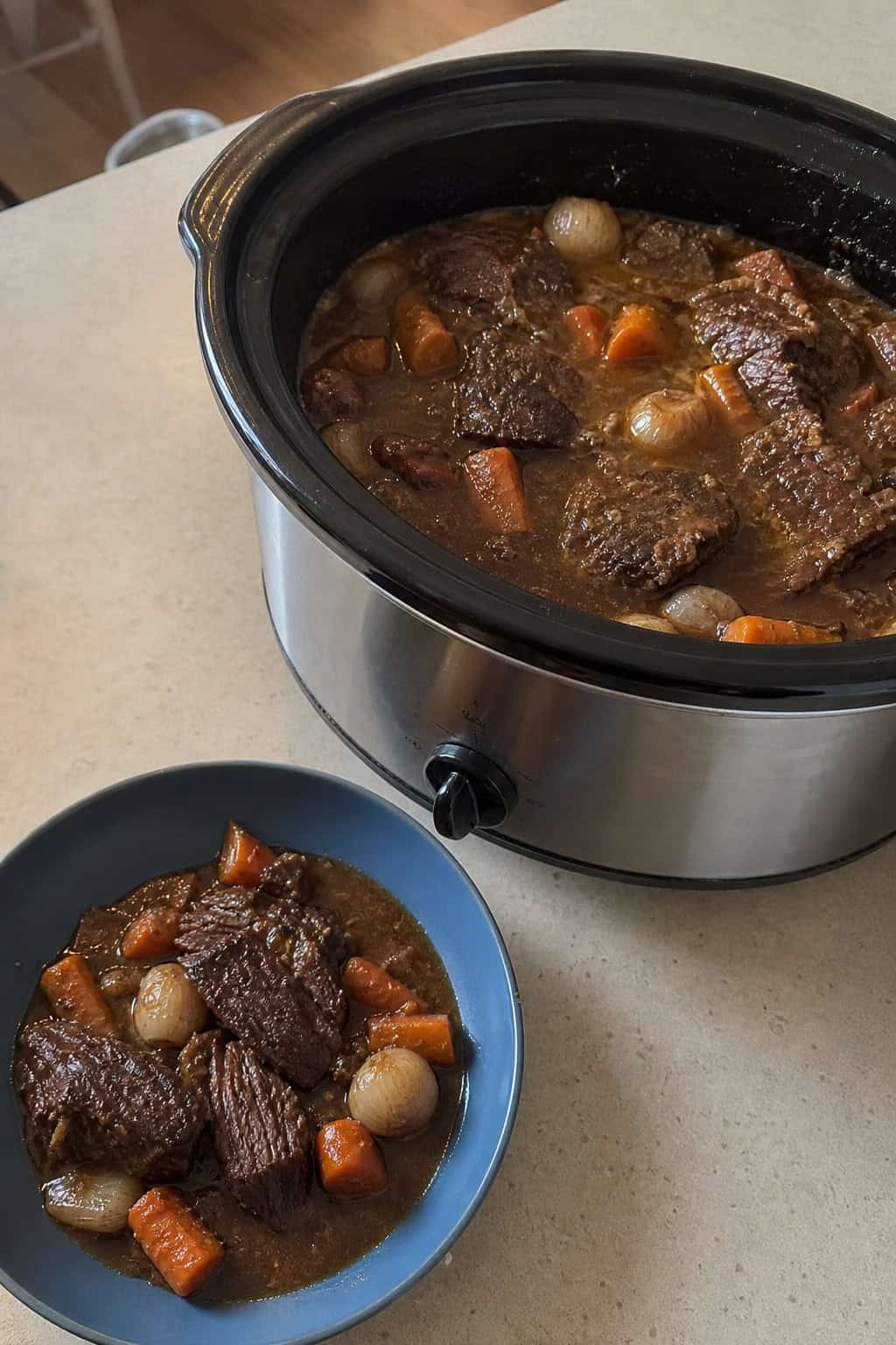 A crockpot filled with beef short rib and red wine stew on a wooden countertop surrounded by fresh vegetables and herbs, with a glass of red wine nearby.