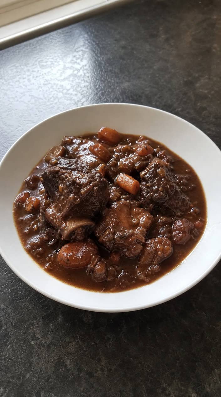 Slow cooker filled with beef short ribs in red wine sauce, garnished with thyme, on a wooden table with cooking ingredients around.