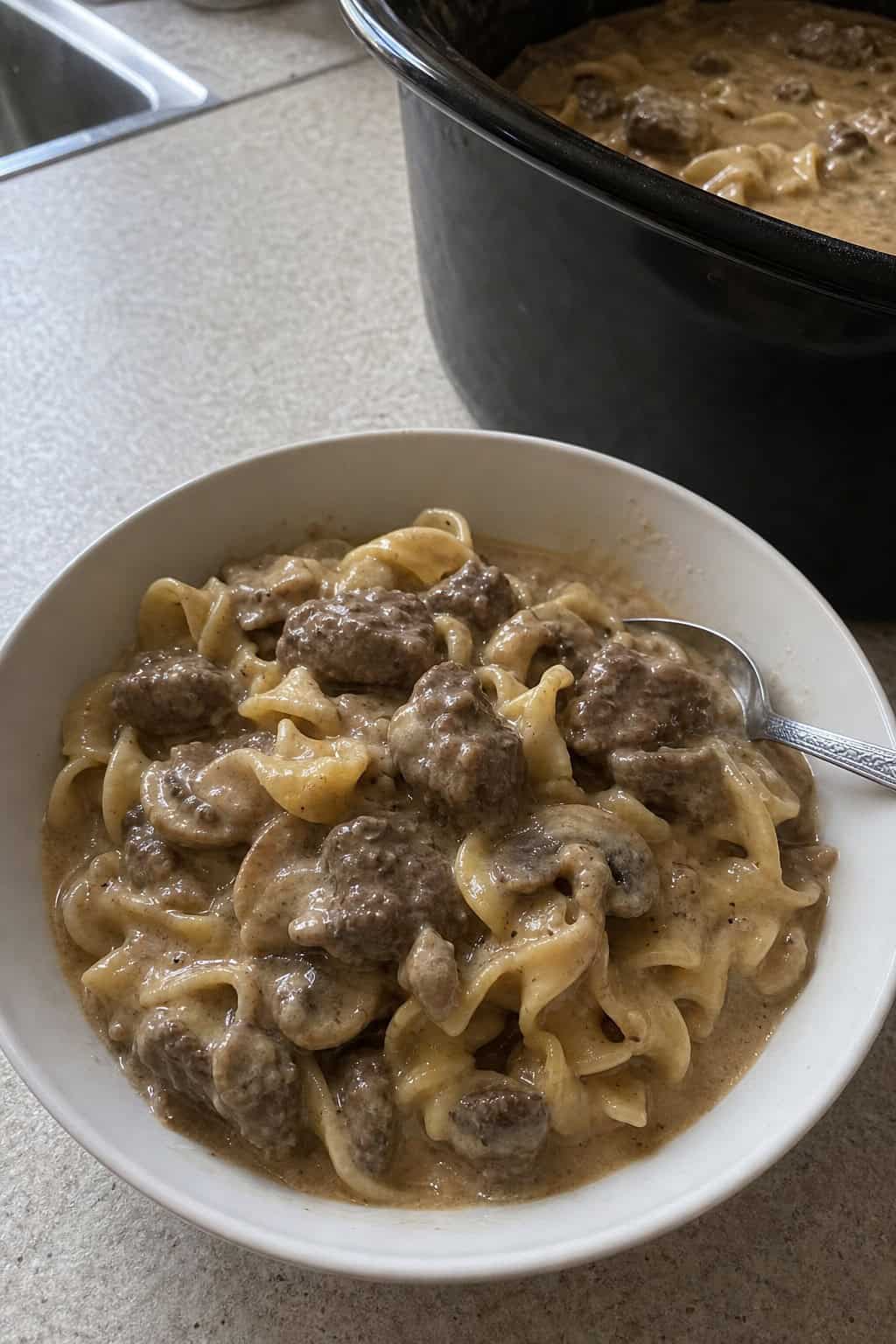 A bowl of beef stroganoff with noodles and mushrooms on a wooden table next to a slow cooker and kitchen utensils.