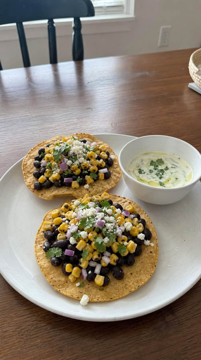 Close-up of black bean and corn tostadas topped with fresh ingredients and a bowl of lime crema on a wooden table.