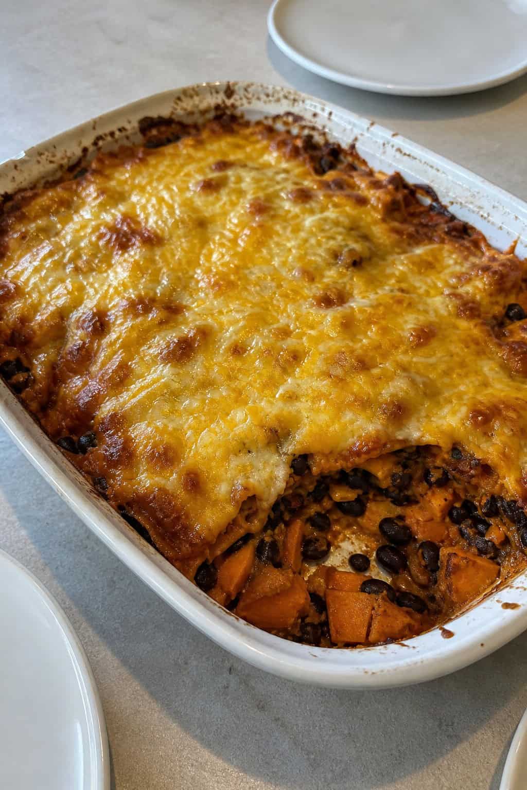 A baked black bean and sweet potato enchilada casserole in a terracotta-style ceramic dish, with visible orange sweet potato chunks and black beans beneath the browned cheese, with lime wedges and fresh cilantro alongside.