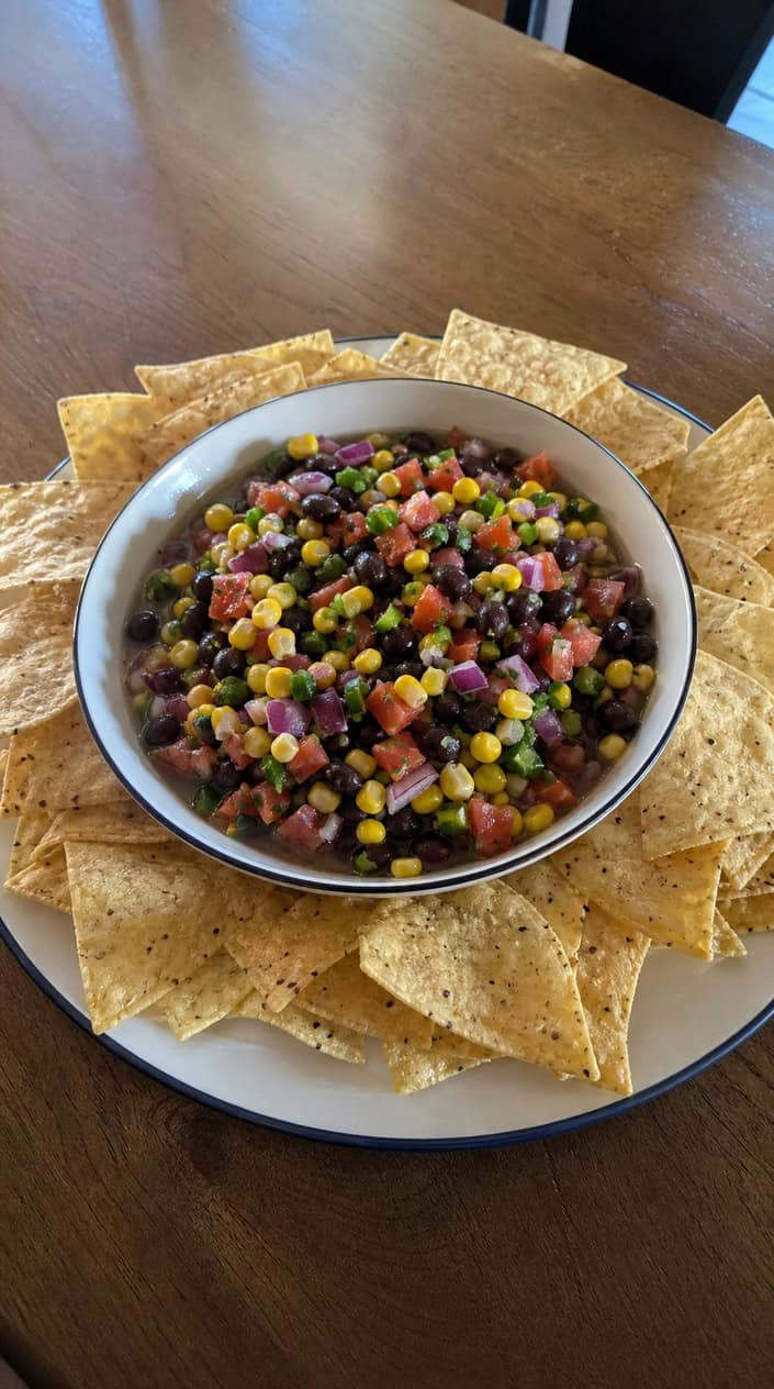 A bowl of black bean and corn salsa with tortilla chips on a wooden table in an outdoor backyard setting.