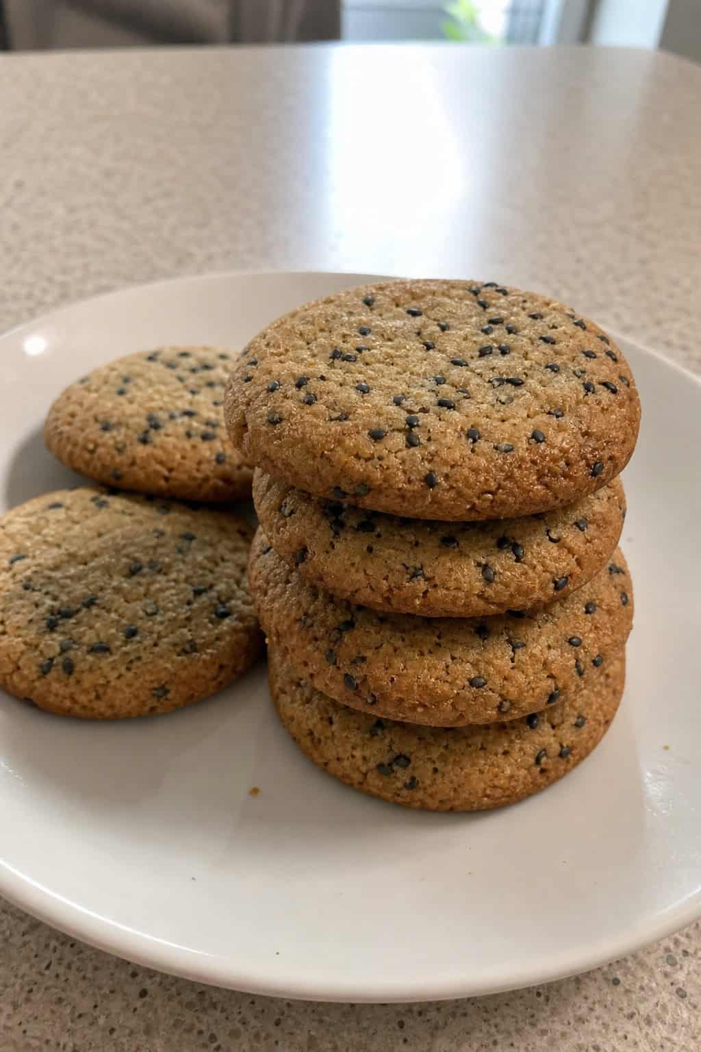 A stack of black sesame brown butter cookies on a wooden surface with scattered sesame seeds and a bowl of melted brown butter nearby.