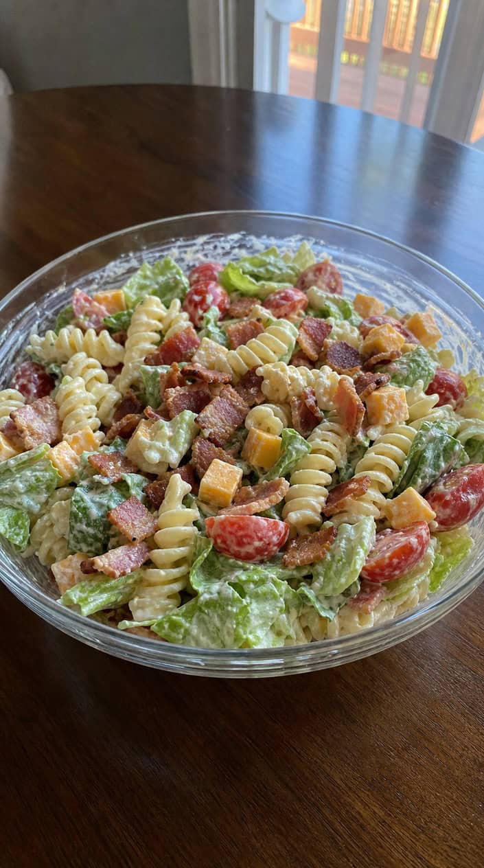 A bowl of BLT pasta salad with bacon, cherry tomatoes, lettuce, and creamy dressing on a wooden table.