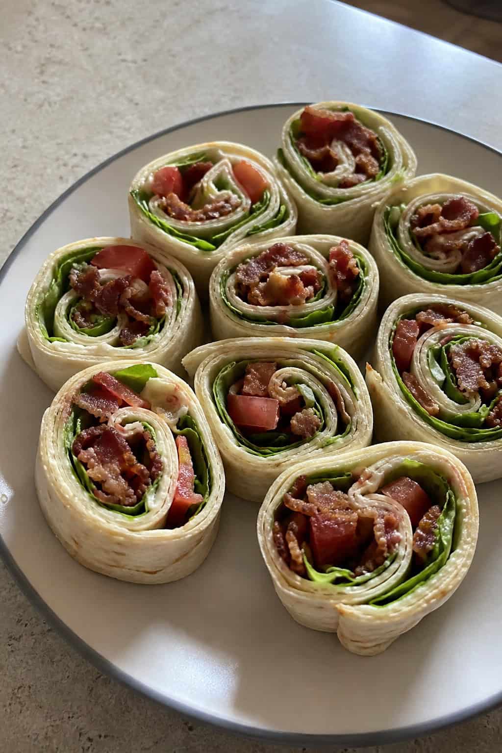 Plate of BLT pinwheel appetizers showing distinct layers of chopped crispy bacon, shredded romaine, and diced tomato inside sliced flour tortilla rolls on a wooden table.