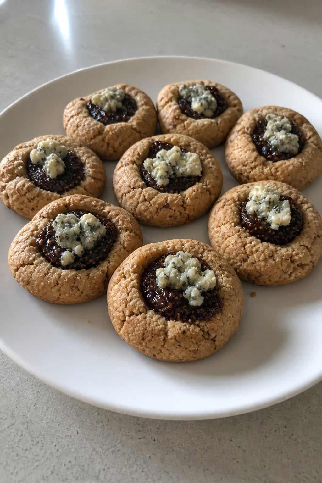 A wooden board with golden brown thumbprint cookies filled with blue cheese and fig jam, surrounded by fresh figs and blue cheese wedges.