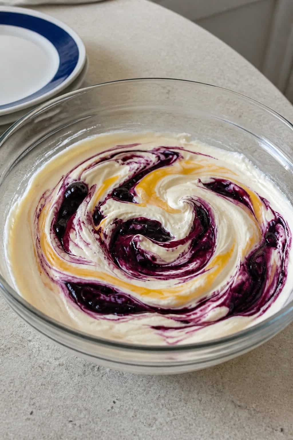 A bowl of creamy cheesecake filling swirled with blueberry and lemon curd, surrounded by fresh blueberries and lemon slices on a kitchen counter.