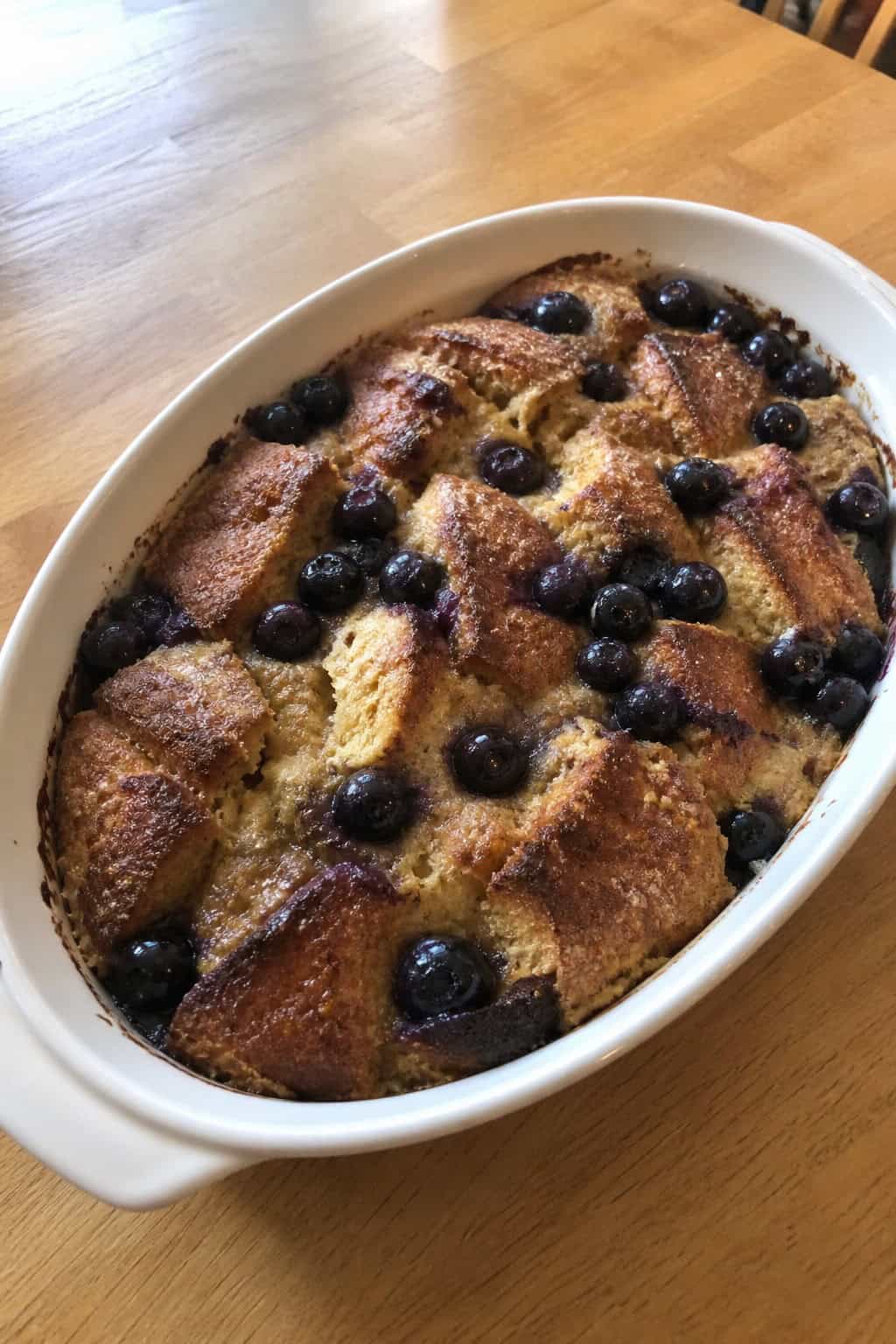 A freshly baked blueberry-lemon French toast casserole in a white ceramic dish on a wooden table with fresh blueberries, a lemon wedge, and a fork placed nearby.
