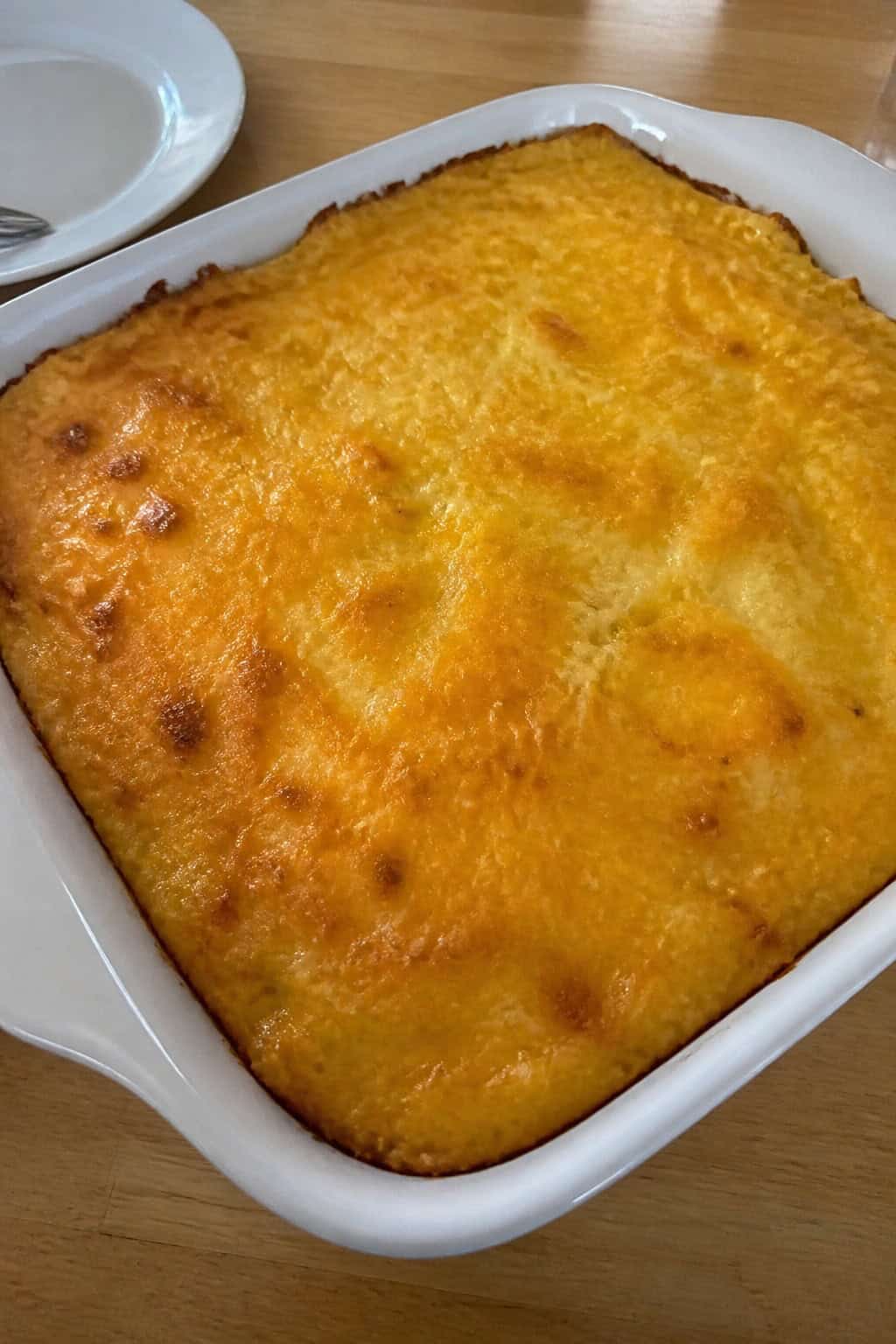 A golden brown hashbrown casserole with melted cheddar cheese and eggs in a baking dish on a wooden table with herbs and a glass of orange juice nearby.