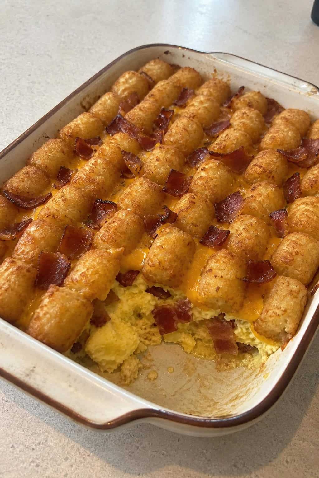 A golden brown breakfast tater tot casserole in a ceramic baking dish, with crispy bacon pieces and set egg custard visible between the tots, just pulled from the oven.