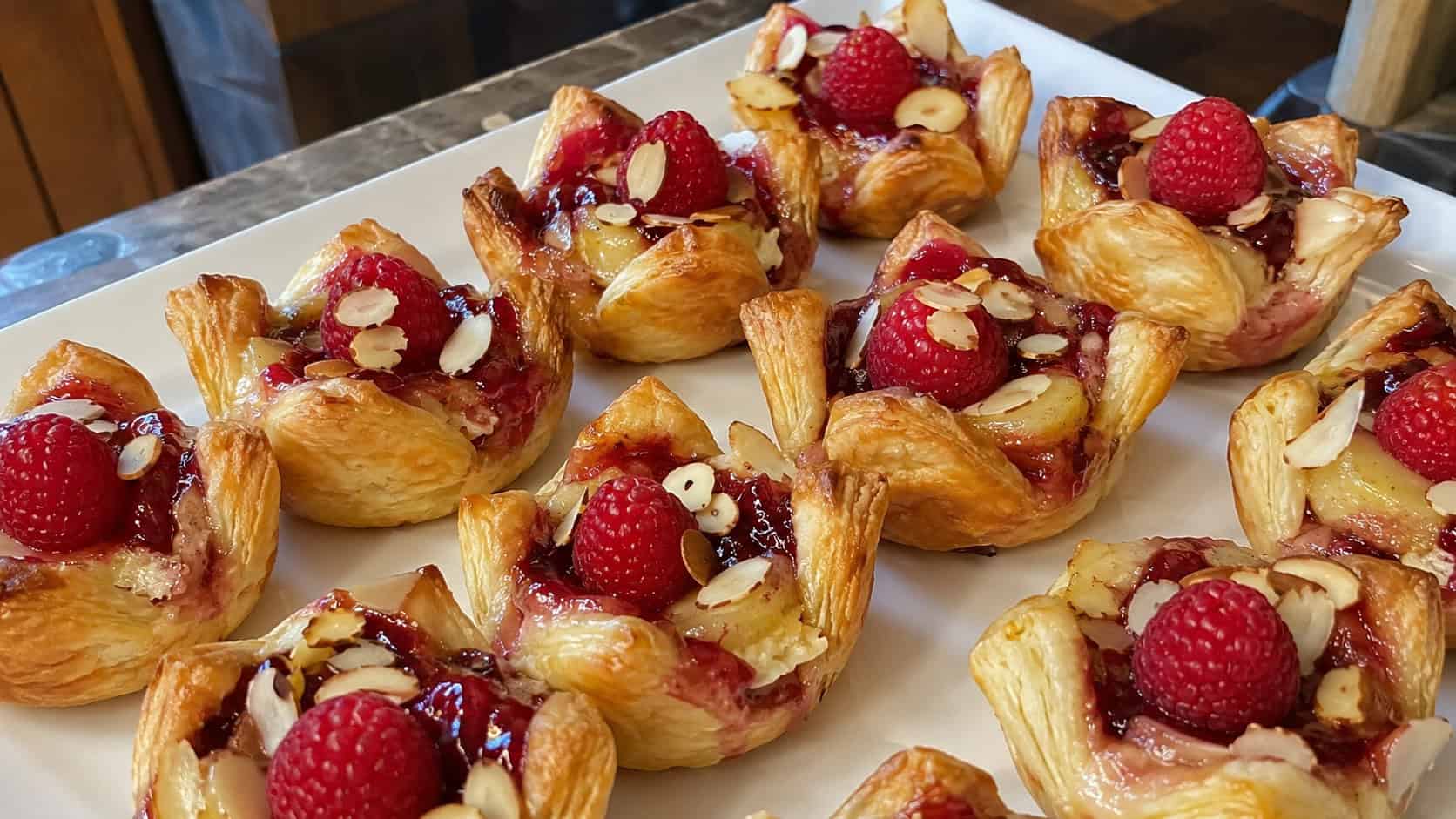 finished brie and raspberry puff pastry bites on a serving plate, golden puffed pastry cups with melted brie and red raspberry jam, a fresh raspberry and sliced almonds on top of each, warm natural light, casual home entertaining