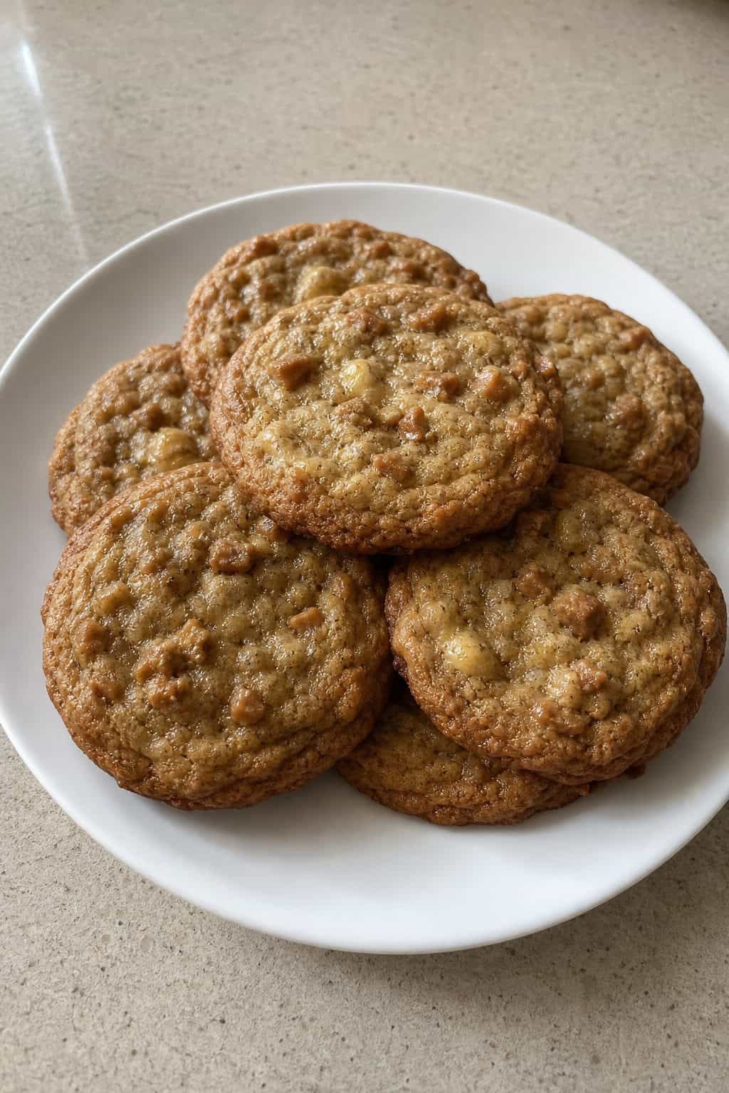 A plate of golden browned-butter banana toffee cookies with ripe bananas and toffee pieces on a wooden table.