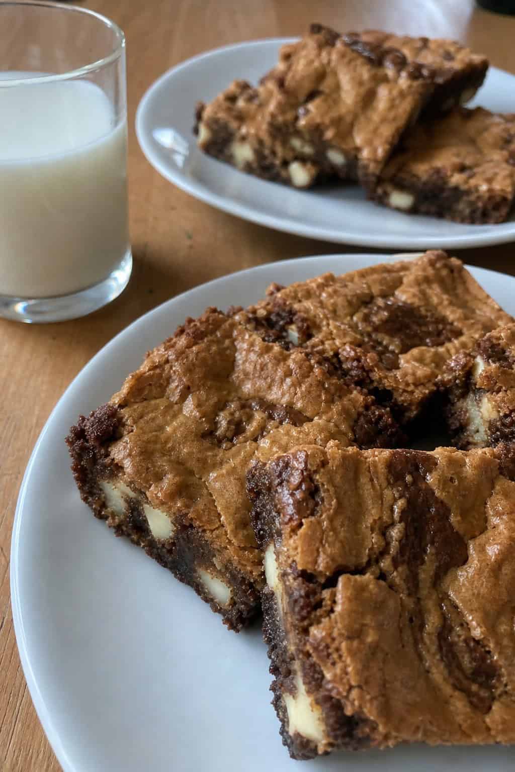Square pieces of brownie-blondie dessert with visible white chocolate chunks and brown sugar on a wooden table, with a bowl of brown sugar and white chocolate chips nearby.
