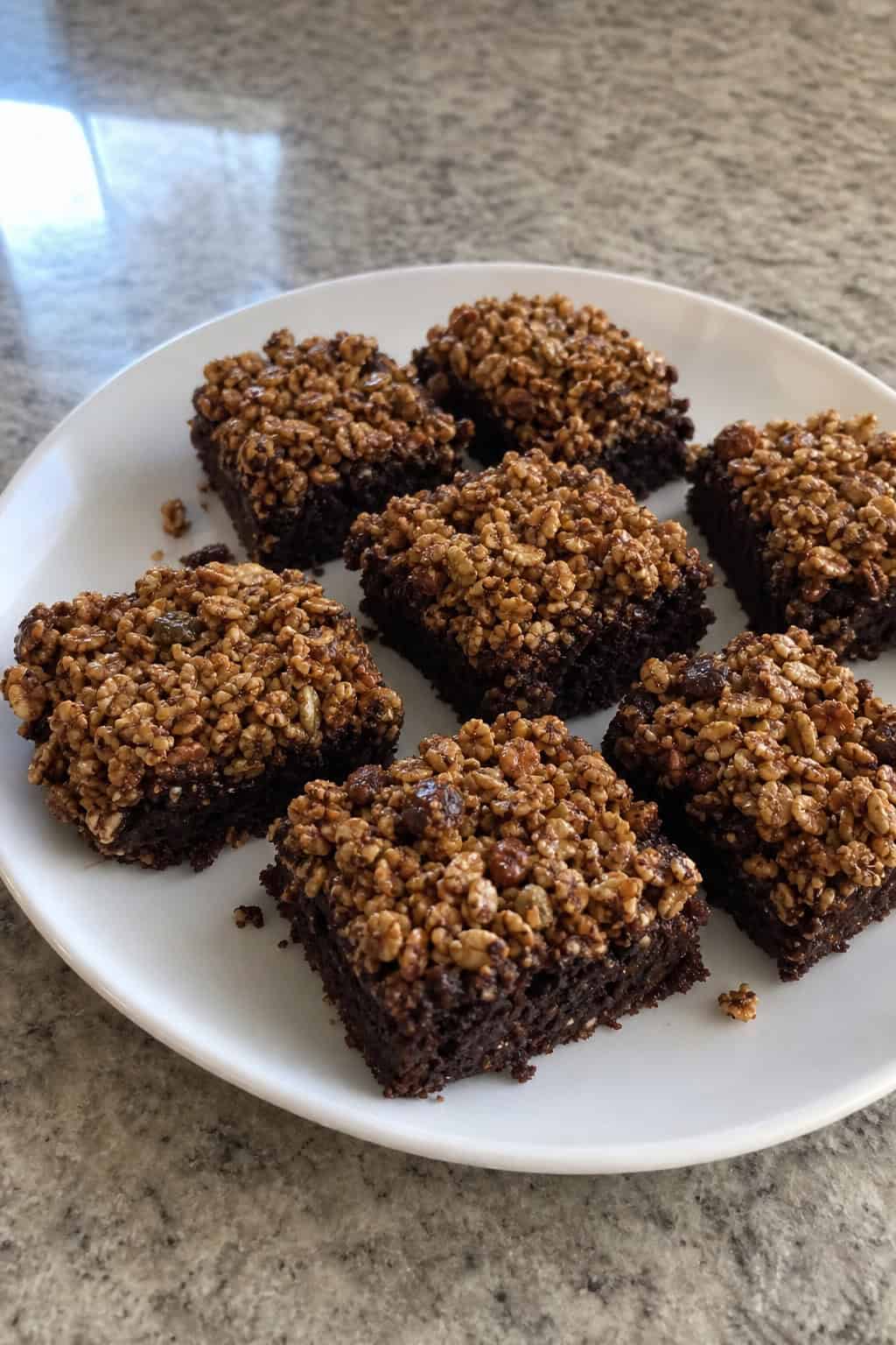 A close-up of square brownie bars topped with granola arranged on a wooden board with a bowl of granola nearby in a kitchen setting.