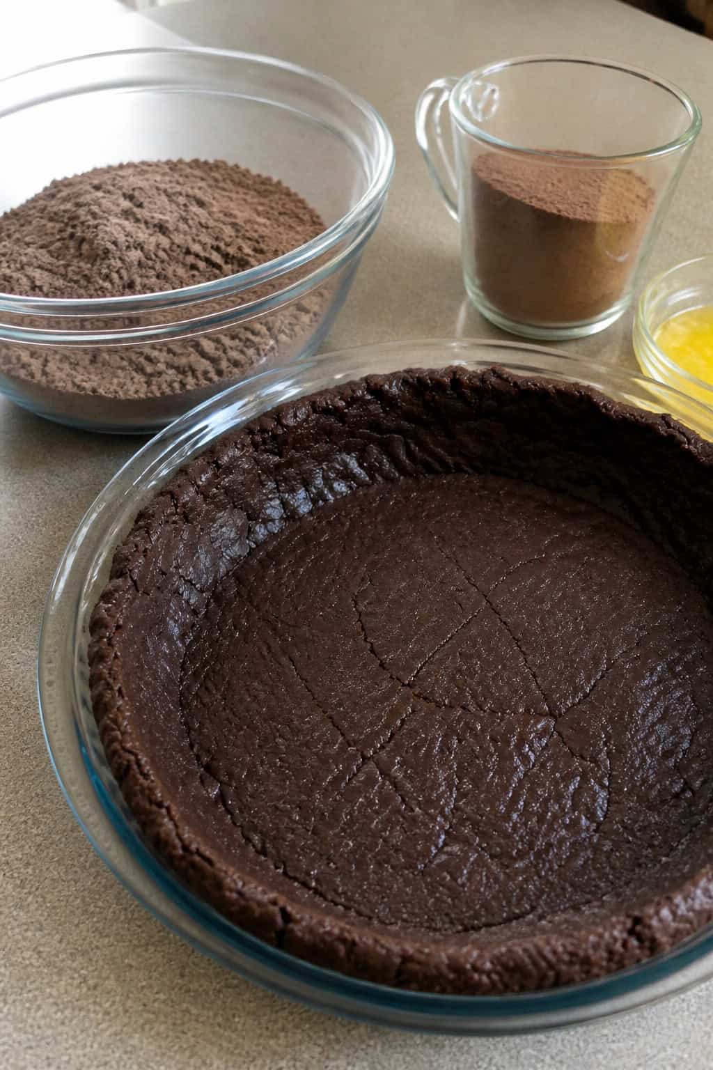 A chocolate brownie mix pie crust in a glass pie dish surrounded by baking ingredients on a wooden countertop.