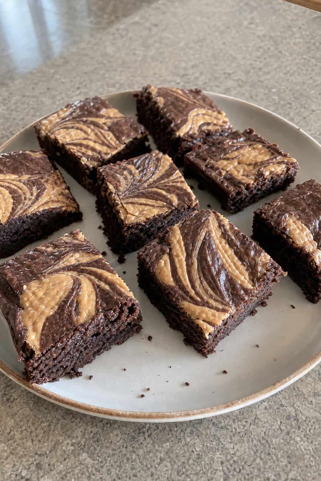 Close-up of square brownie bars with peanut butter swirls on a wooden board with baking ingredients in the background.
