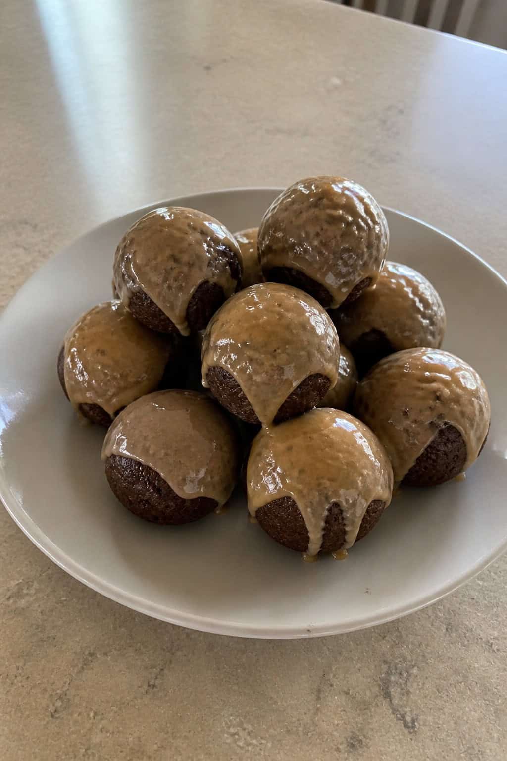 Plate of small brownie donut holes glazed with coffee icing on a wooden table with coffee beans and a cup of coffee in the background.