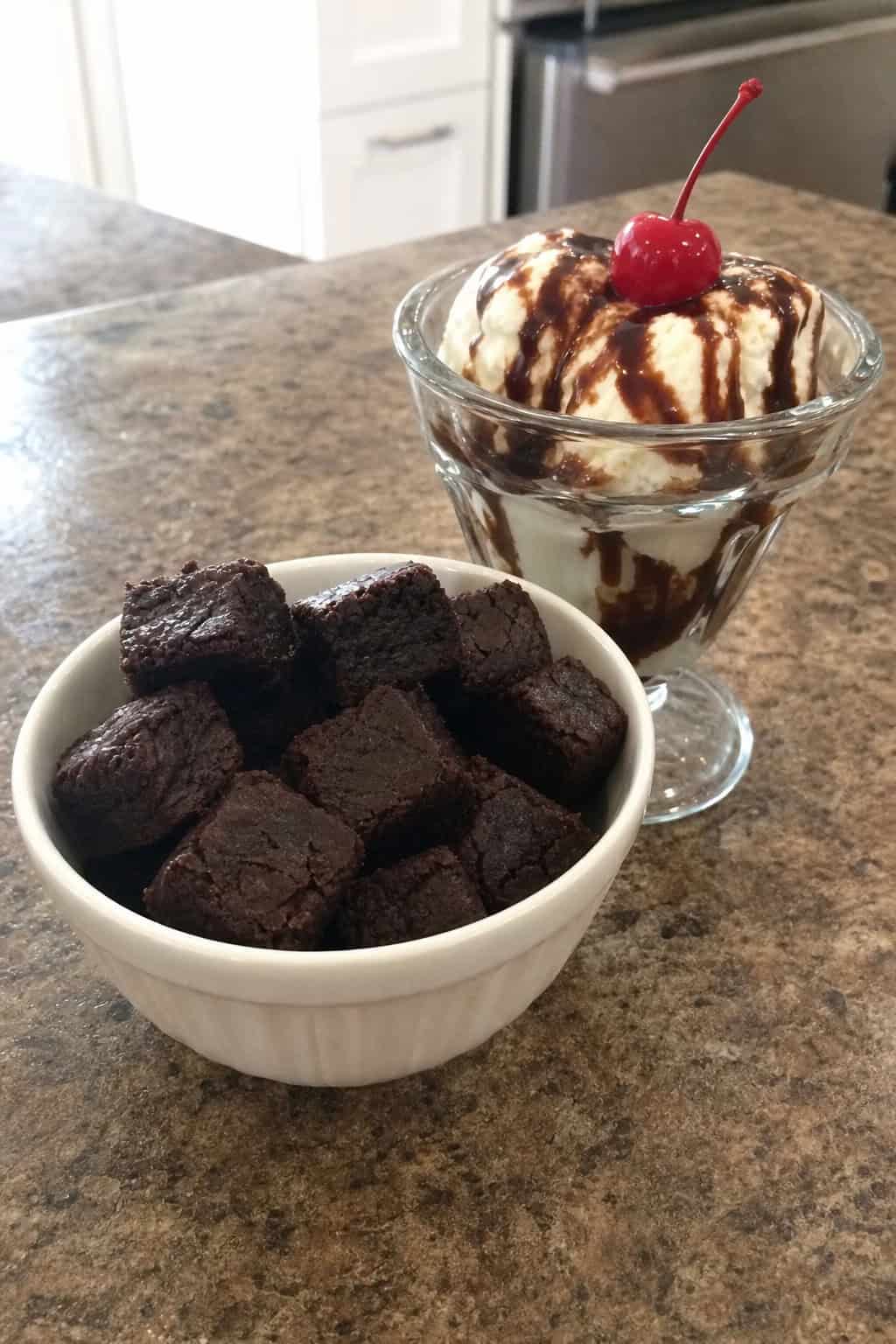 A bowl of fudgy brownie croutons next to an ice cream sundae topped with chocolate syrup and a cherry on a wooden surface.